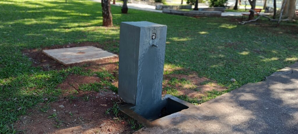 Esta imagem mostra um bebedouro público de concreto, pintado de cinza, posicionado em um espaço ao ar livre, provavelmente um parque ou praça. Aqui estão os detalhes que observo: O Bebedouro: É uma coluna retangular simples de concreto. Na parte superior, há um botão de acionamento e uma pequena torneira ou bico de saída de água. É possível notar que ele está um pouco úmido, sugerindo que foi usado recentemente. A Base: Na base do bebedouro, há uma pequena caixa ou ralo embutido no chão, desenhado para coletar a água que escorre da torneira e evitar que ela se acumule ao redor. O Ambiente: O bebedouro está situado na transição entre uma área gramada e um caminho ou calçada de concreto (à direita). Ao fundo, o parque parece bem cuidado, com gramado verde, algumas árvores cujas sombras se projetam no chão, e o que parece ser um banco de praça à distância. Iluminação: A luz solar está forte, criando sombras bem definidas, o que indica um dia ensolarado e claro.
