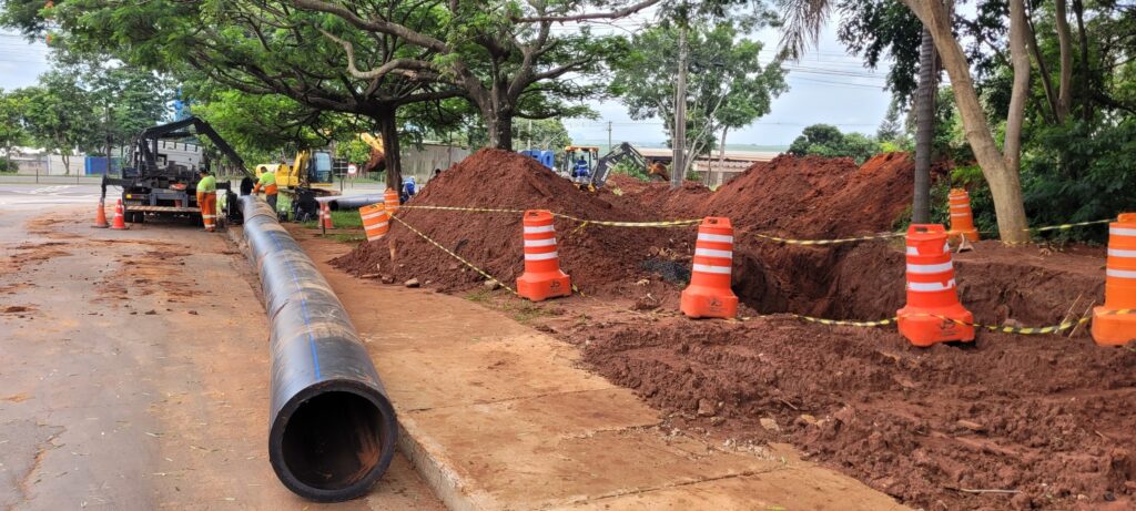 Esta imagem mostra uma obra em andamento, provavelmente de instalação de rede de água ou esgoto, em uma área urbana. Aqui estão os detalhes: Tubulação: Há um longo tubo preto de grande diâmetro estendido ao longo da calçada. Escavação: Ao lado do tubo, há uma vala aberta e grandes montes de terra vermelha que foram escavados. Equipamento: É possível ver um caminhão com guindaste à esquerda, ajudando no manuseio do tubo, e uma retroescavadeira mais ao fundo, entre os montes de terra. Segurança: A área está sinalizada com cones laranja e faixas de sinalização zebradas (amarelo e preto) para garantir a segurança dos pedestres e trabalhadores. Pessoal: Alguns trabalhadores uniformizados (com coletes reflexivos) estão presentes no local supervisionando ou executando a tarefa. Parece ser uma obra de infraestrutura típica, bem sinalizada e em execução durante o dia.