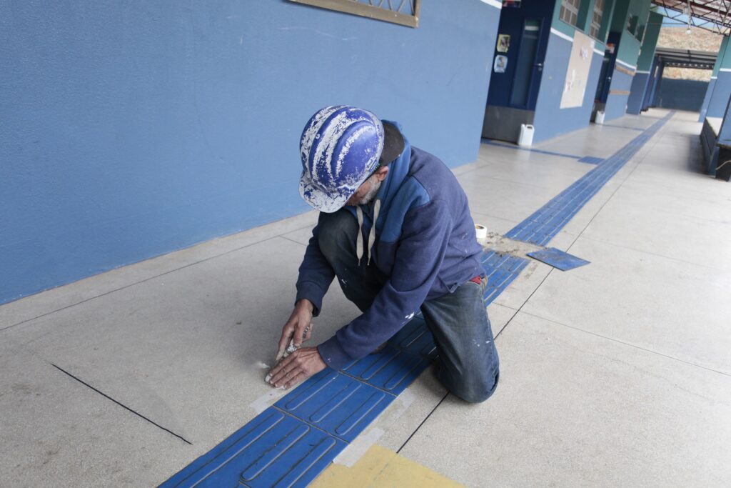 Um trabalhador agachado, visto de perfil/atrás, instalando ou fazendo acabamento em uma faixa tátil azul no piso de um corredor coberto. Pessoa: usa capacete azul com respingos de tinta branca, moletom/agulha azul e calça jeans; cabelos/barba visíveis mas sem identificação. Ação: mãos próximas ao piso aplicando massa/cola ou alisando o rejunte sobre as placas táteis azuis (linha guia para pessoas com deficiência visual). Piso e sinalização: faixa tátil azul contínua ao longo do corredor; também há uma marca amarela quadrada próxima. Ambiente: corredor amplo e coberto com paredes pintadas de azul, portas e murais ao fundo — parece ser área escolar ou prédio público. Ferramentas/insumos: pequeno rolo/recipiente e restos de material de assentamento ao lado do trabalhador. Iluminação e clima: dia claro com luz natural entrando pelas laterais; cena transmite trabalho manual cuidadoso e manutenção acessível.