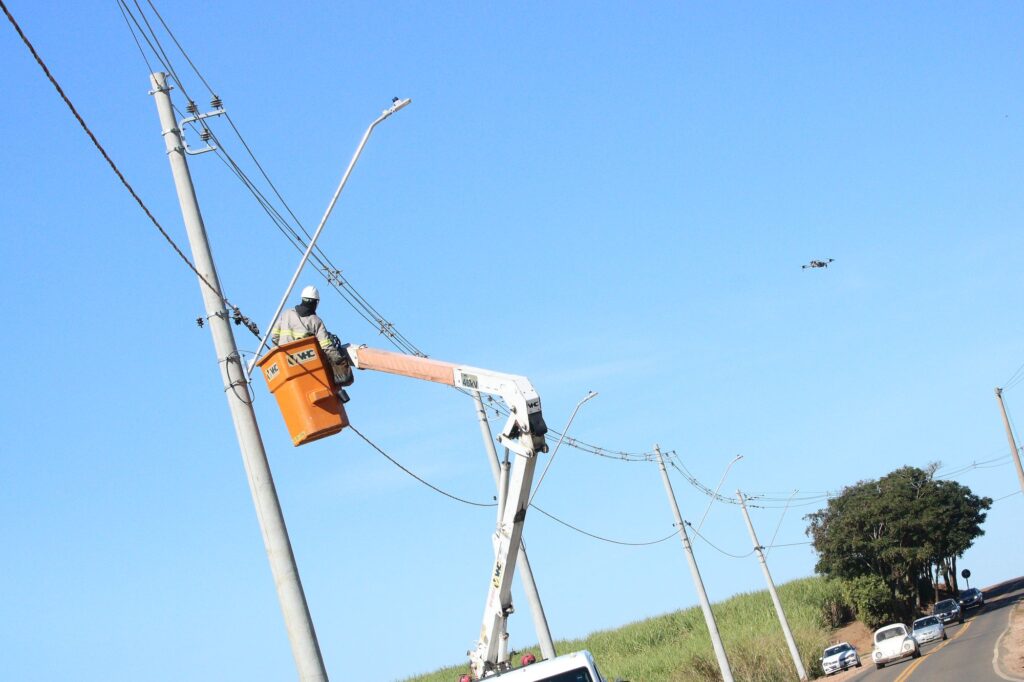 A imagem mostra uma equipe de manutenção elétrica trabalhando em uma área rural. Um caminhão com plataforma elevatória está estacionado ao lado de uma estrada asfaltada, e um trabalhador, usando equipamentos de segurança, realiza ajustes em um poste de energia. Outro profissional opera o sistema no solo.