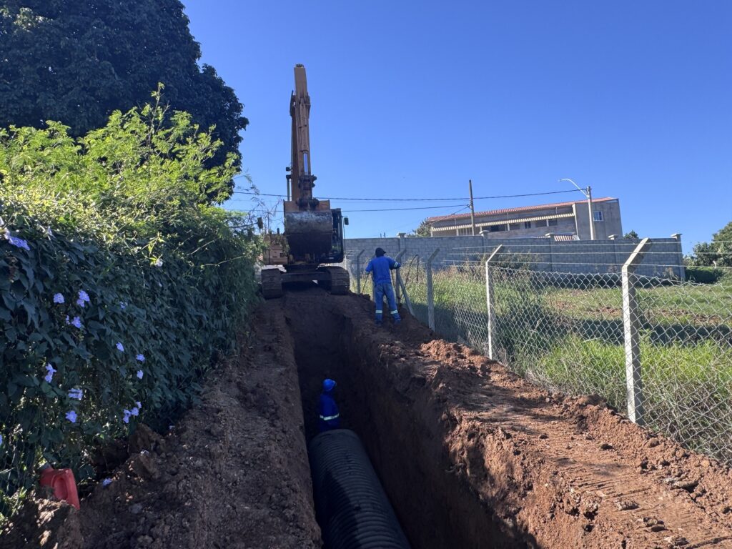 Foto colorida de uma obra externa durante o dia: escavação de uma vala profunda com colocação de uma tubulação corrugada preta. Maquinaria e trabalhadores: Uma escavadeira grande, posicionada no topo da vala, com o braço e a caçamba voltados para frente. Dois trabalhadores com roupa azul (um dentro da vala, outro na borda) — não é possível identificar quem são. Vala e tubulação: Vala estreita e profunda ao longo do trajeto da obra; no fundo já há uma tubulação corrugada preta assentada. Solo avermelhado retirado e acumulado nas bordas, com marcas das esteiras da máquina. Ambiente e limites: À direita, cerca de arame e postes de concreto separando o canteiro de um terreno gramado e de um prédio ao fundo. À esquerda, vegetação densa com flores roxas e uma grande árvore que projeta sombra. Céu e iluminação: Céu azul limpo, sol forte e sombras nítidas, indicando manhã ou início de tarde. Impressão geral: Trabalho de infraestrutura (provavelmente drenagem ou escoamento pluvial) em andamento, organizado e com uso de equipamentos pesados.