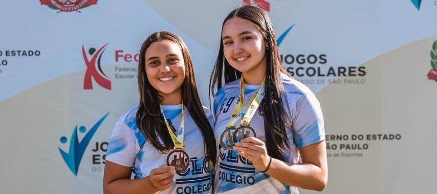 Foto de duas jovens posando sorrindo com medalhas em um pódio/área de premiação. Principais elementos: Pessoas: duas meninas adolescentes, lado a lado, sorrindo para a câmera; ambas seguram medalhas com fita amarela e detalhes em verde (provavelmente medalhas de participação/pódio). Roupa: camisetas esportivas azul-claro com padrão em tons de azul e a palavra “COLÉGIO” visível na parte frontal — indicam participação escolar. Acessórios: cada uma usa uma medalha pendurada no pescoço; uma delas segura a medalha mais à frente para exibir. Fundo: painel de divulgação com logotipos — lê-se “Jogos Escolares”, “Governo do Estado de São Paulo” e símbolos da federação/organização do evento, confirmando contexto esportivo/escolar. Atmosfera: momento de comemoração e orgulho, luz natural suave e ótima expressão facial que transmite felicidade e conquista. Detalhes menores: cabelo solto em ambas; numerais de identificação no ombro de uma camiseta (ex.: “19”); cores limpas e composição centralizada.