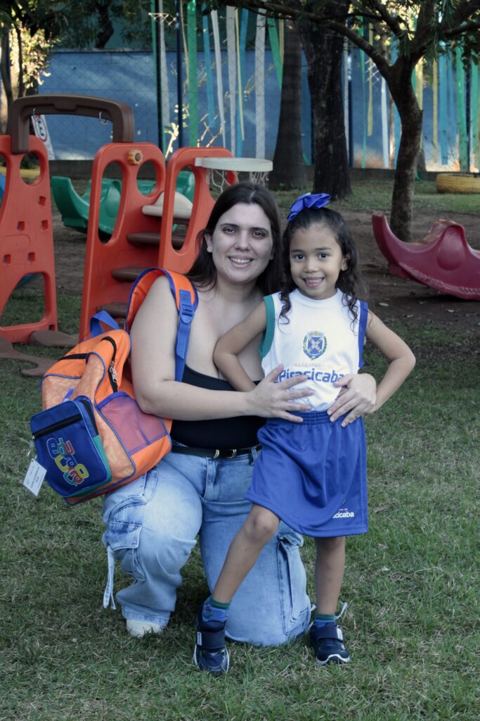 Uma mulher agachada abraça uma menina pequena em um parquinho ao ar livre — ambas sorriem para a câmera. Posição e expressão: a mulher está agachada no joelho esquerdo, com o braço envolvendo a menina; as duas sorriem, olhar direcionado à câmera. Roupa e acessórios: a menina usa uniforme escolar branco e azul com o brasão e a palavra “Piracicaba” no peito, saia/short azul, meias verdes e tênis escuro; tem um laço roxo no cabelo. A mulher veste regata preta e jeans claros rasgados; carrega uma mochila escolar laranja e azul apoiada no ombro. Cenário: parquinho com brinquedos plásticos coloridos (vermelho e laranja) e piso de grama; árvores e uma cerca ao fundo, tiras decorativas verdes penduradas mais ao fundo. Detalhes: a menina apoia a mão na cintura da mulher; a mochila tem etiqueta e estampa colorida “educa…” visível; luz natural suave, ambiente de fim de tarde ou manhã clara. Atmosfera: cena afetuosa e casual, provavelmente momento de chegada ou saída da escola, transmite cuidado e alegria entre adulta e criança.