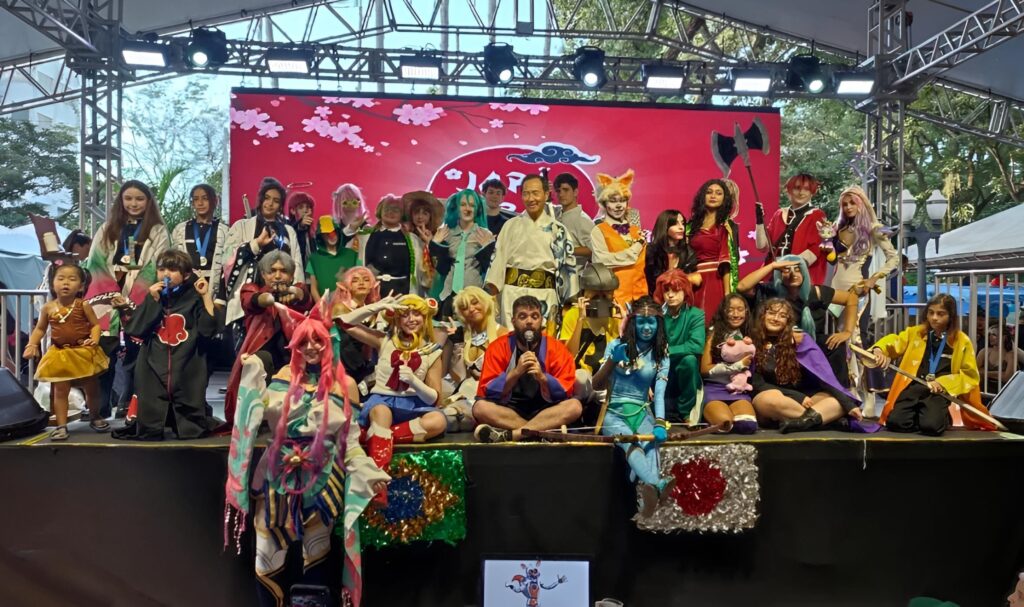 grupo grande de cosplayers posando no palco durante um evento ao ar livre — foto colorida, cheia de fantasias variadas e personagens, com painel vermelho decorativo ao fundo. Pontos principais: Cena: palco coberto com iluminação de evento e telão/painel vermelho ao fundo com elementos florais; ambiente externo (árvores visíveis atrás). Pessoas: cerca de 25–30 participantes de idades variadas em poses de grupo — muitos jovens e crianças; ninguém é identificado. Fantasias: diversidade grande de personagens (cores vibrantes, peles pintadas, perucas, armaduras, adereços como machados, espadas e elementos mangá/anime/games). Atitude: clima festivo e de celebração — alguns estão sentados à frente, outros em pé atrás, sorrisos e poses de cosplay tradicionais. Objetos e cenário: decoração de palco, estrutura metálica de iluminação, placas e enfeites na frente do palco; público e barracas do evento visíveis lateralmente. Impressão geral: apresentação ou concurso de cosplay em convenção/feira, com energia alegre, variedade criativa e forte presença visual.