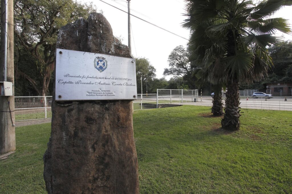 Esta é uma foto de um monumento histórico em um espaço ao ar livre. O Monumento: Trata-se de uma grande pedra bruta que serve de base para uma placa comemorativa. A Placa: Na placa está escrito que "Piracicaba foi fundada neste local em 01/08/1767 por Capitão Povoador Antônio Corrêa Barbosa". Abaixo, menciona o "223º Aniversário de Fundação" e a "Prefeitura Municipal de Piracicaba". O Cenário: O monumento está posicionado em uma área gramada. Ao lado, há palmeiras e, ao fundo, percebe-se uma área pavimentada com uma grade de proteção, postes de luz e alguns carros estacionados, sugerindo ser um espaço público ou parque urbano.