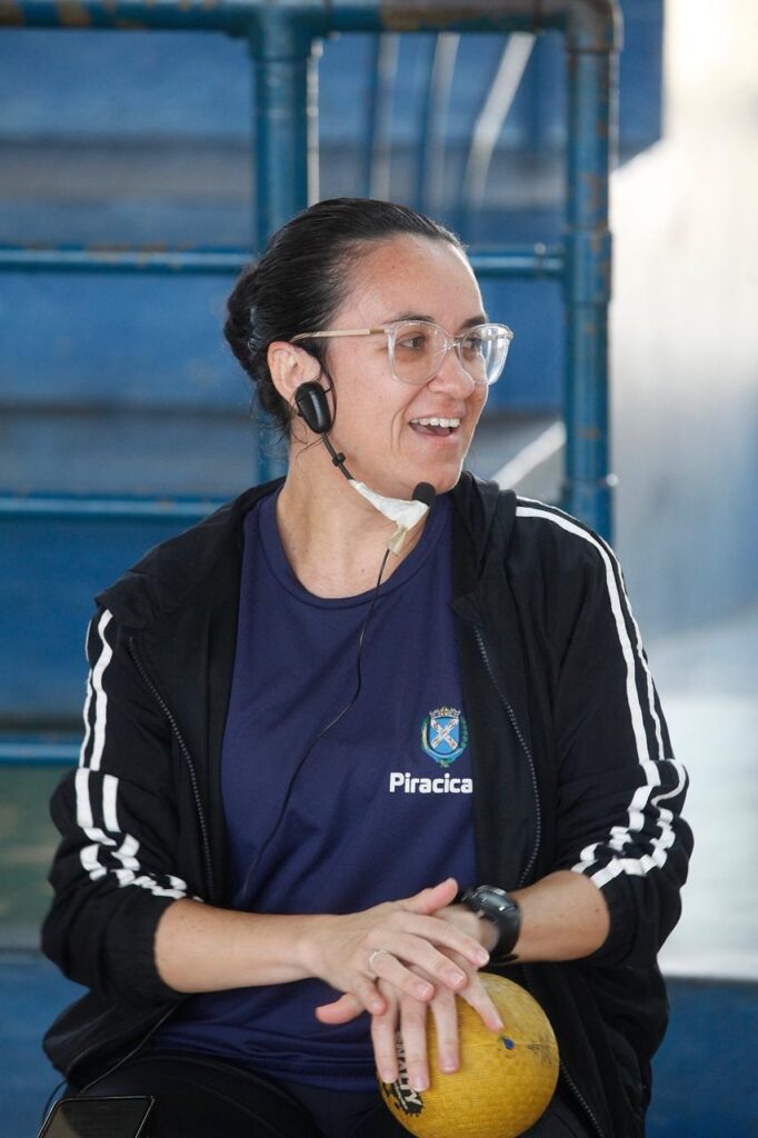 Mulher sentada em ambiente de quadra ou ginásio, segurando uma bola amarela com as duas mãos apoiadas sobre ela. Atmosfera casual e de atividade física; parece estar em intervalo ou conversa com o grupo. Pessoas e expressão Mulher de meia‑idade, cabelos escuros presos em coque, usando óculos de armação transparente. Sorri levemente e olha para a direita, expressão aberta e comunicativa. Roupas e acessórios Veste camiseta azul-marinho com o brasão e a palavra “Piracicaba” impressos no peito. Jaqueta preta com listras brancas nas mangas. Usa headset com microfone preso próximo à boca; o microfone está fixado com fita branca na parte inferior do queixo. Relógio escuro no pulso esquerdo e aliança discreta na mão esquerda.