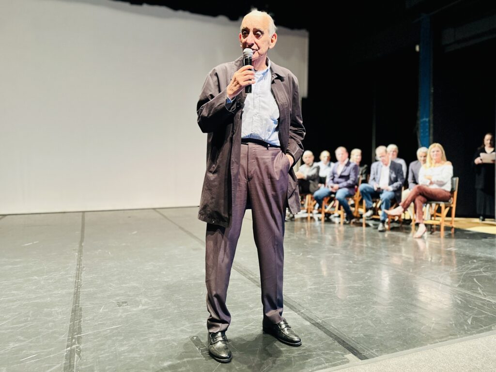 Foto de um orador em um palco durante um evento institucional, com público/mesa de painel ao fundo. Observações principais: Pessoa em primeiro plano: homem adulto idoso segurando um microfone com a mão direita e gesticulando levemente com a esquerda no bolso; veste paletó/jaqueta escura sobre camisa clara e calça social marrom/acinzentada. Postura e expressão: em pé, postura relaxada e confiante, olhando para a plateia; não faço identificação pessoal. Palco e cenário: cenário escuro atrás do orador com uma grande tela branca/clair à esquerda (provavelmente usada para projeção). Piso do palco em primeiro plano mostra marcas e linhas típicas de teatro/ auditório. Público/painel: ao fundo, à direita, um grupo de cerca de oito pessoas sentadas em cadeiras enfileiradas — alguns observam o orador, outros parecem atentos; figurinos formais e semiformal (blazers, camisas, calças). Uma mulher em primeiro plano à direita veste blusa branca e calça marrom. Iluminação: foco de luz intenso sobre o orador, criando contraste entre ele e o fundo escuro; iluminação mais suave sobre as pessoas sentadas ao fundo. Composição/fotografia: enquadramento vertical que privilegia o orador em plano próximo, com profundidade mostrando o painel ao fundo; sensação de evento formal — palestra, lançamento ou apresentação. Atmosfera: institucional e séria, com tom de discurso público ou apresentação técnica.