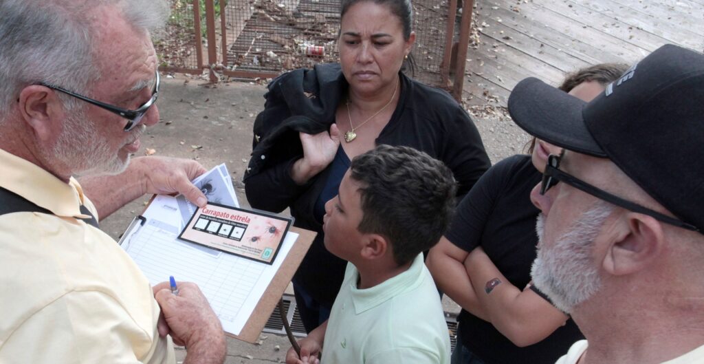 Cena externa de orientação/registro sobre segurança em saúde (cartilha sobre “carrapato estrela”). Resumo: um homem mais velho, à esquerda, segura uma prancheta com um formulário e folhetos; ele escreve com uma caneta enquanto mostra um cartão informativo sobre “carrapato estrela”. Pessoas: ao centro, uma mulher de expressão séria/atenta segura uma jaqueta sobre o ombro e observa o material; na frente dela, um menino olha para o cartão; à direita aparece outro homem de barba e boné escuro, também prestando atenção. Há pelo menos mais uma pessoa parcialmente visível atrás. Roupas e acessórios: o homem com a prancheta usa óculos e camisa clara; a mulher veste roupa escura e um colar com pingente em forma de coração; o menino usa polo clara; o homem de boné usa óculos escuros presos ao boné. Material informativo: um folheto/cartão com imagens de carrapatos e texto em português (“carrapato estrela”) e um formulário em anexo na prancheta. Ambiente: área externa com piso de concreto, madeira e uma grade metálica ao fundo; folhas secas no chão indicam espaço ao ar livre, possivelmente próximo a vegetação. Tom da cena: informativo e sério — parece uma ação de conscientização ou coleta de dados sobre prevenção/risco de picadas de carrapato.