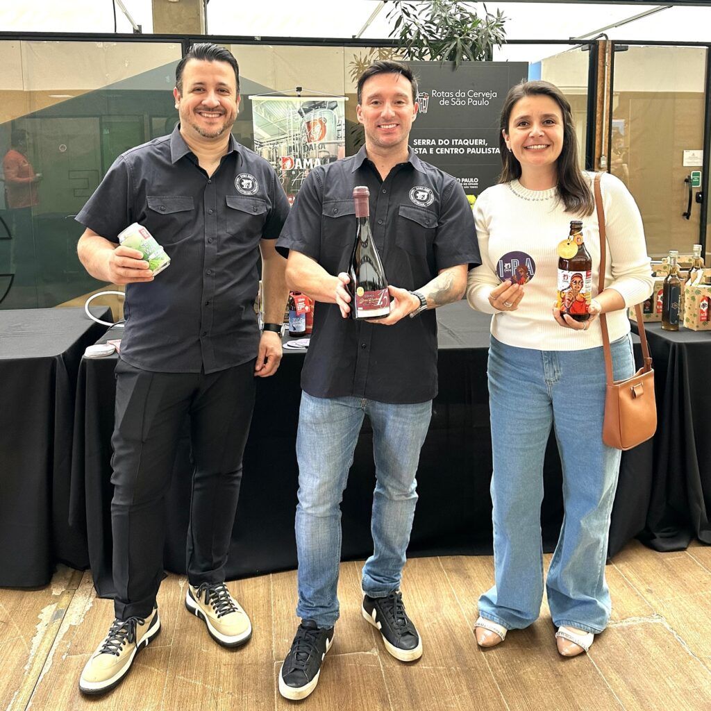 Três pessoas posam lado a lado em um estande/mesa de exposição, sorrindo e mostrando produtos (garrafas e latas) — parece um evento de cervejarias ou feira gastronômica. Ambiente: estande com painel ao fundo (há um banner com imagem de equipamento de cervejaria e texto promocional parcialmente visível). Mesa coberta por toalha preta e algumas embalagens ao redor. Pessoas: duas homens à esquerda e uma mulher à direita, em pé, ombros próximos. Homem à esquerda: camisa preta de botão com logo bordado no peito, calça preta e tênis claros; segura uma lata. Homem no centro: também usa camisa preta com logo e jeans; segura uma garrafa grande de vinho/cerveja artesanal com rótulo vermelho. Mulher à direita: blusa clara de mangas longas, jeans azul claro e bolsa tiracolo marrom; segura uma garrafa com rótulo ilustrado e um adesivo/crachá redondo. Objetos e detalhes: garrafas e latas com rótulos coloridos e ilustrados; o rótulo central mostra o número “15”. Piso de madeira; plantas ao fundo; iluminação natural/indoor. Tom geral: foto casual e profissional ao mesmo tempo — parece promoção e degustação de produtos artesanais, com clima amistoso e descontraído.