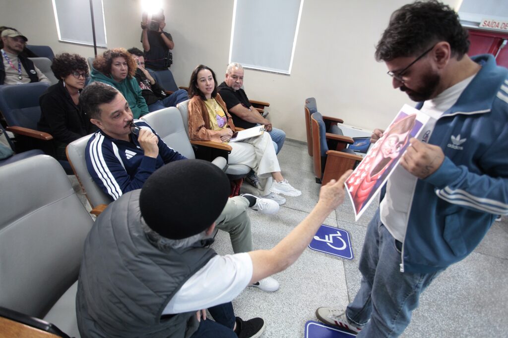 Esta foto mostra um encontro em sala tipo auditório ou sala de atendimento, com várias pessoas sentadas em cadeiras alinhadas prestando atenção a um homem em pé à direita. Cena geral: fileira de cadeiras acolchoadas; piso em granito ou cerâmica; placas de sinalização de acessibilidade no chão. Pessoas sentadas: grupo heterogêneo de adultos (homens e mulheres) em diferentes idades, alguns anotando ou segurando prancheta; expressões atentas e curiosas. Um homem em primeiro plano segura os óculos e parece concentrado. Pessoa em pé: um homem de perfil, usando jaqueta esportiva azul e óculos, segura uma impressão colorida (uma ilustração) que recebe de outra pessoa sentada — parecem avaliar ou comentar a imagem. Atmosfera: clima de reunião, oficina ou avaliação — interação próxima, informal e focada; iluminação interna e janela com cortina ao fundo. Detalhes visuais: alguém ao fundo fotografa com flash apontado para a cena; roupas casuais (moletom, jaqueta, tênis); cadeiras com apoios de madeira; parede clara com placa de saída visível no canto superior direito. A imagem transmite um momento de discussão participativa, provavelmente relacionado a apresentação ou análise de um material visual.