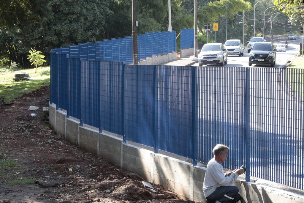 A imagem mostra um operário trabalhando na instalação ou manutenção de uma grade metálica azul, que está sendo fixada sobre um muro de concreto. Ele está agachado na parte externa, trabalhando na base da cerca. Ao fundo, há uma via urbana com carros circulando e uma placa de sinalização de trânsito indicando a presença de ciclistas. O cenário parece ser uma área pública, com uma área gramada e arborizada à esquerda e uma infraestrutura urbana consolidada.