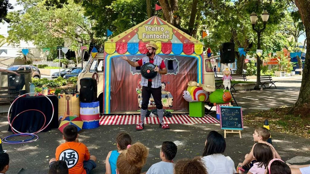 Esta é uma foto encantadora de uma apresentação de circo ao ar livre, voltada para o público infantil. O artista: No centro, um palhaço com nariz vermelho, chapéu de palha e meias listradas está performando, segurando um objeto redondo na mão e fazendo um gesto como se contasse uma história. O cenário: A apresentação acontece em frente a uma Kombi que foi adaptada como um "Teatro de Fantoche". O veículo está decorado com cores vivas (amarelo, vermelho e azul), criando uma atmosfera mágica e lúdica. Detalhes: Ao lado do palhaço, há decorações temáticas como um caracol grande e colorido. Há uma pequena lousa à direita avisando "Kombi no Circo - 10h". À esquerda, vemos um suporte com bambolês e um alto-falante. O público: Um grupo de crianças está sentado no chão, de costas para a câmera, assistindo atentamente à performance em uma praça arborizada e ensolarada.
