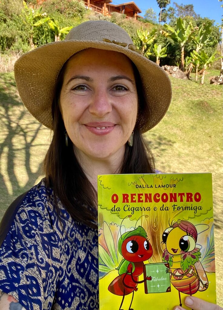 Mulher sorrindo em close segurando um livro infantil à frente do peito. Aparência: rosto em close, pele clara, sorriso suave, cabelos castanhos longos à mostra. Usa brincos alongados e um chapéu de palha de aba larga. Roupa: blusa com estampa azul e bege em padrão geométrico/fluido. Livro: capa colorida com o título "O REENCONTRO da Cigarra e da Formiga" (autora: Dalila Lamour). Ilustração mostra uma formiga vermelha e uma cigarra amarela, estilo cartunesco, segurando um livro verde. Ambiente: área externa ensolarada — gramado, sombras de árvores ao fundo, plantas (bananeiras) e algumas casas de madeira no topo de um pequeno morro. Iluminação e clima: luz natural forte, dia claro com céu azul aparente no canto superior; sombras suaves no rosto e no chão.