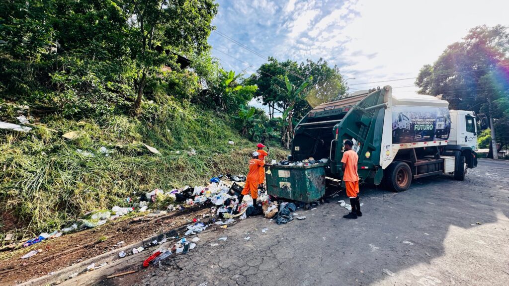 A imagem mostra três garis uniformizados com roupas laranjas trabalhando na coleta de lixo em uma via pública. Dois deles estão manipulando uma grande caçamba de lixo metálica para despejar o conteúdo na parte traseira de um caminhão de lixo verde. O caminhão possui uma plotagem lateral com a mensagem "Coletando o presente e preservando o futuro" e o nome da empresa "Lopac". Há uma quantidade significativa de lixo espalhado no chão, próximo a uma encosta gramada com vegetação e algumas bananeiras. O dia parece ensolarado, com um céu azul e algumas nuvens.