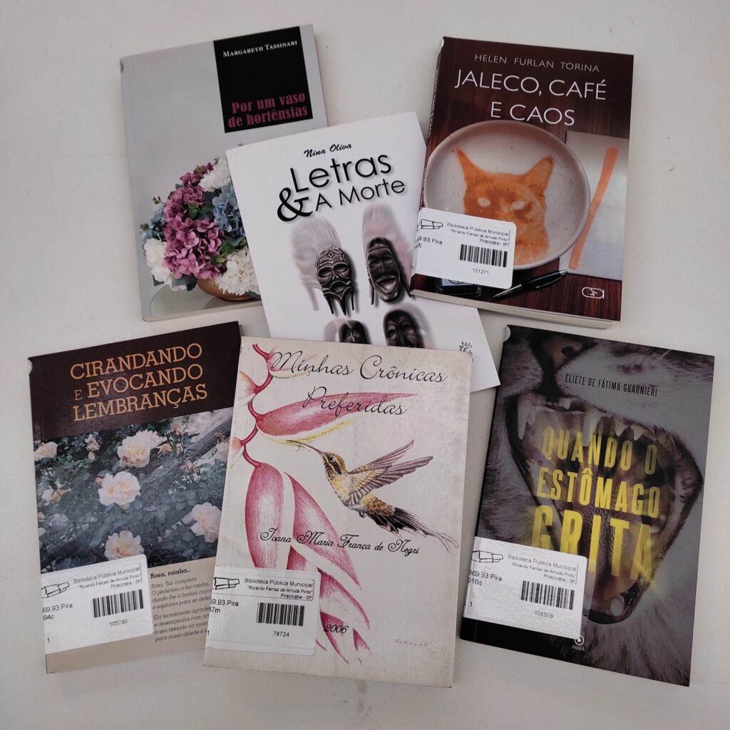 A imagem mostra seis livros dispostos sobre uma superfície clara. Pelo que observei, eles parecem pertencer à Biblioteca Pública Municipal "Ricardo Ferraz de Arruda Pinto", em Piracicaba, já que alguns possuem etiquetas de identificação da biblioteca. Aqui estão os títulos que aparecem: Por um vaso de hortênsias, de Margareth Tassinari. Letras & A Morte, de Nina Oliva. Jaleco, Café e Caos, de Helen Furlan Torina. Cirandando e Evocando Lembranças, cuja capa mostra flores brancas. Minhas Crônicas Preferidas, de Ivana Maria França de Negri (com um beija-flor na capa). Quando o Estômago Grita, de Eliete de Fátima Guarnieri (com a imagem de uma boca aberta na capa).
