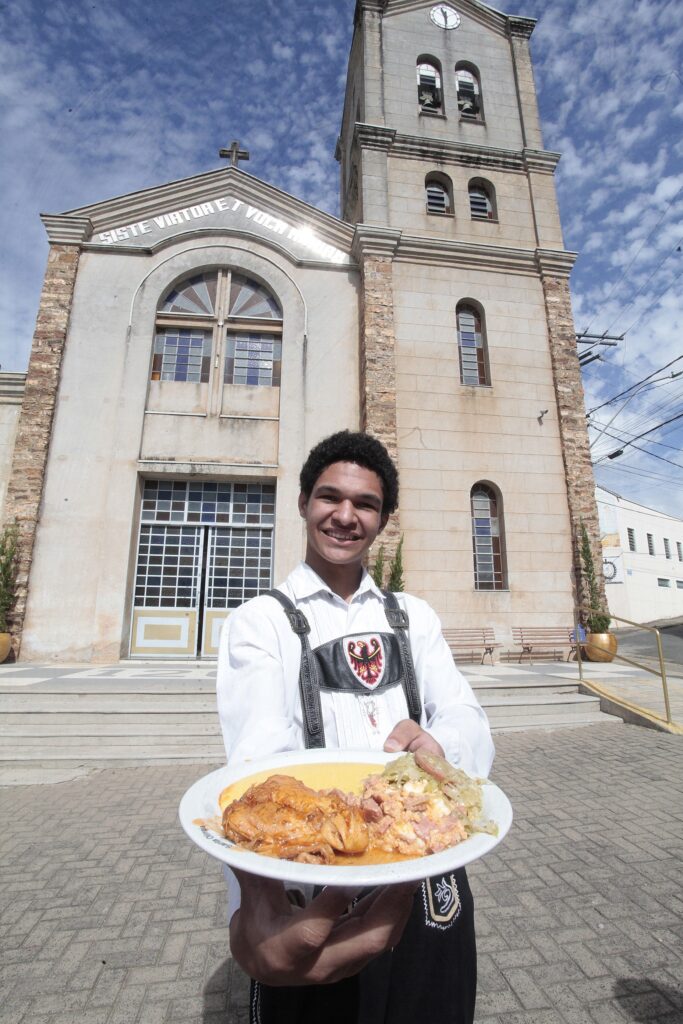 Foto de uma pessoa adulta jovem em frente a uma igreja, segurando um prato com comida e sorrindo para a câmera. Observações detalhadas: Pessoa: jovem, sorriso aberto, pele de tom médio-escuro, cabelo curto e cacheado. Veste uma camisa branca e um traje com peitoral decorado (parece um traje folclórico/cerimonial com bordado ou brasão no centro). Usa mochila de alças visíveis sobre os ombros. Ação: estende um prato branco com comida em direção à câmera — prato contém pedaços de frango ao molho e acompanhamentos (aparenta ser uma mistura de arroz, farofa/creme salgado e legumes refogados). Há vapor leve indicando a comida quente. Ambiente/arquitetura: igreja em estilo clássico atrás da pessoa, com fachada de pedra clara, torre sineira à direita com relógio e sinos visíveis. Acima da entrada há um frontão com inscrição em letras grandes (parece texto em latim). Portas e janelas com grades e vitrais simples. Degraus levam à entrada; bancos e vasos decorativos aparecem na lateral. Iluminação e clima: dia ensolarado com céu parcialmente nublado (muitas nuvens pequenas), luz forte que projeta sombras definidas. A foto tem perspectiva levemente de baixo para cima (ângulo que valoriza a torre da igreja). Composição e sensação: foco humano em primeiro plano (sorriso e prato) contra cenário religioso/urbano ao fundo — transmite hospitalidade, celebração local ou tradição comunitária (possivelmente festa, missa com almoço, quermesse ou evento cultural). Ambiente limpo e organizado; presença de fiação elétrica visível ao fundo à direita, sugerindo área urbana. Detalhes observáveis: ornamento no peitoral do traje com desenho lembrando uma águia ou brasão; calçamento em blocos/pavers na praça; corrimão metálico ao lado dos degraus.