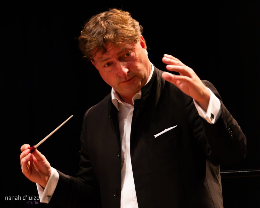 A foto mostra um maestro em pleno momento de condução. Ele está vestindo um terno preto elegante com camisa branca, segurando uma batuta na mão direita, enquanto faz um gesto expressivo com a mão esquerda. O fundo é totalmente escuro, o que destaca ainda mais a figura do maestro e a seriedade da sua concentração. No canto inferior esquerdo, há uma marca d'água discreta indicando "nanah d'luize studio".