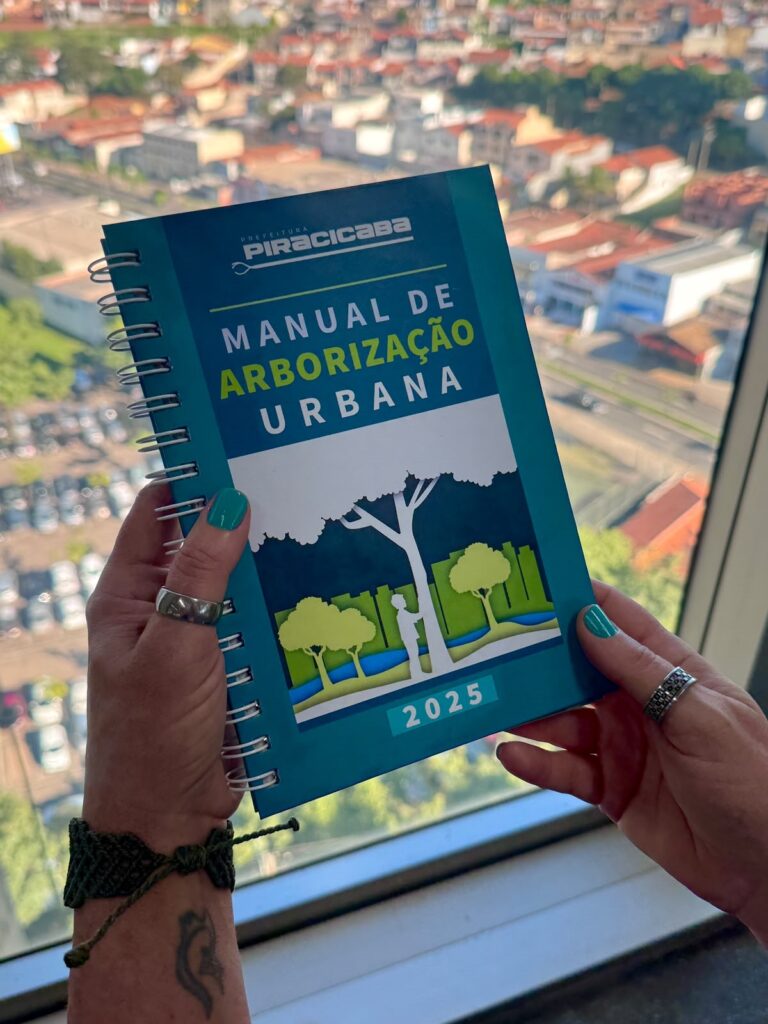 Um caderno espiral nas mãos de uma pessoa, mostrado em primeiro plano diante de uma vista urbana desfocada pela janela. Capa azul e turquesa com ilustração de árvores e silhuetas; título em letras grandes: "MANUAL DE ARBORIZAÇÃO URBANA" com destaque em verde para "ARBORIZAÇÃO" e, no topo, o selo/texto "PREFEITURA PIRACICABA". Na base aparece o ano "2025". Detalhes das mãos e acessórios: Mãos femininas segurando o caderno pelas bordas esquerda e direita. Unhas pintadas em tom verde-azulado combinando com a capa. Anéis prateados nos dedos: um anel largo na mão esquerda e dois anéis na mão direita (um estilo aliança com detalhes). Pulseira trançada verde-escura no pulso esquerdo e uma pequena tatuagem preta visível no antebraço (padrão curvo/ornamental). Ambiente: Ao fundo, desfocado, vê‑se uma cidade com casas e prédios de telhados avermelhados, ruas e áreas verdes — a janela indica que a foto foi tirada em andar alto. Luz natural forte ilumina a cena, realçando as cores vivas da capa e o brilho nas unhas e nos metais dos anéis.