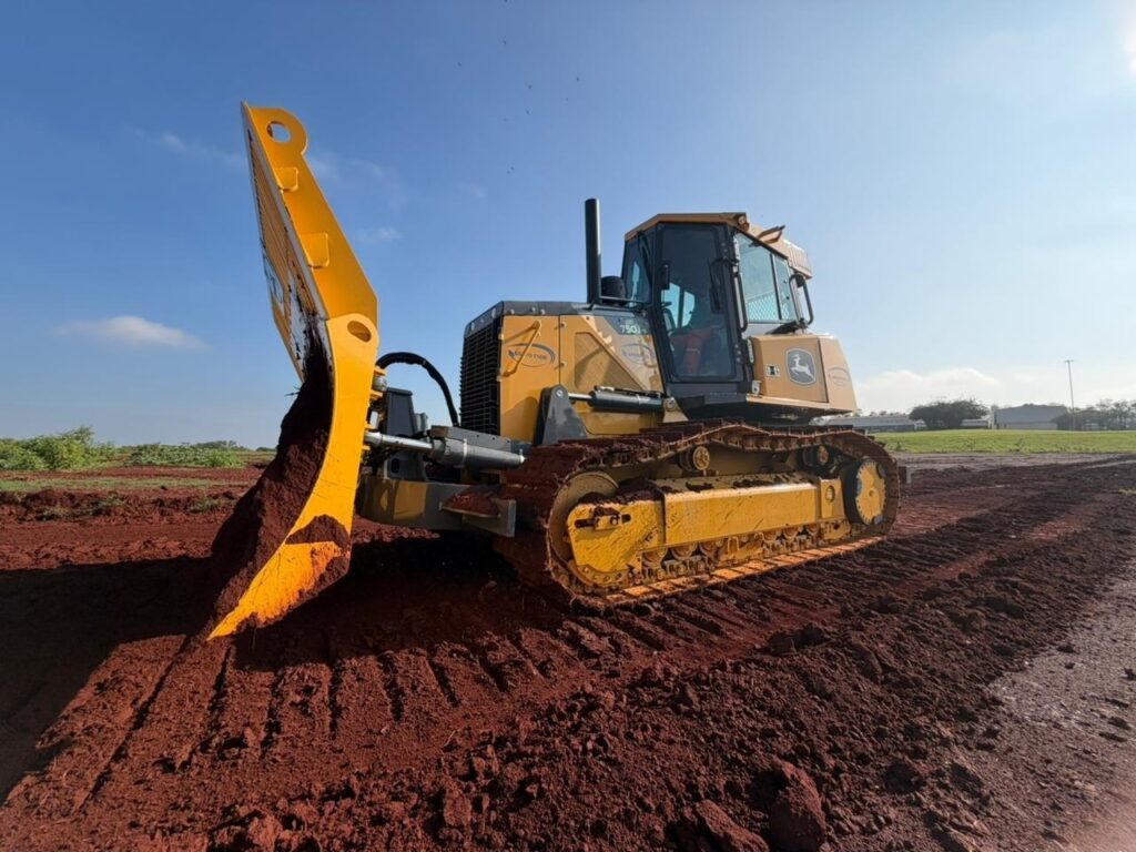 Foto de uma máquina pesada (trator de esteiras / bulldozer) operando em um terreno de terra escura. Máquina: trator de esteiras amarelo com cabine fechada; logotipo visível que parece ser da John Deere na lateral. Ação: lâmina frontal inclinada empurra uma grande quantidade de terra/solo escuro. Posição/ângulo: vista lateral-direita, máquina ocupando a parte direita da imagem; lâmina e monte de terra na parte central-baixa. Ambiente: terreno areno-argiloso com sulcos de movimentação; faixa de grama curta à frente; palmeiras alinhadas ao fundo; céu claro e ensolarado, poucas nuvens. Detalhes adicionais: trilhos metálicos com algo de lama/terra; sombras suaves indicam luz do dia; ao fundo, estrutura/letreiro pequeno com texto (difuso) — possivelmente área pública ou parque.