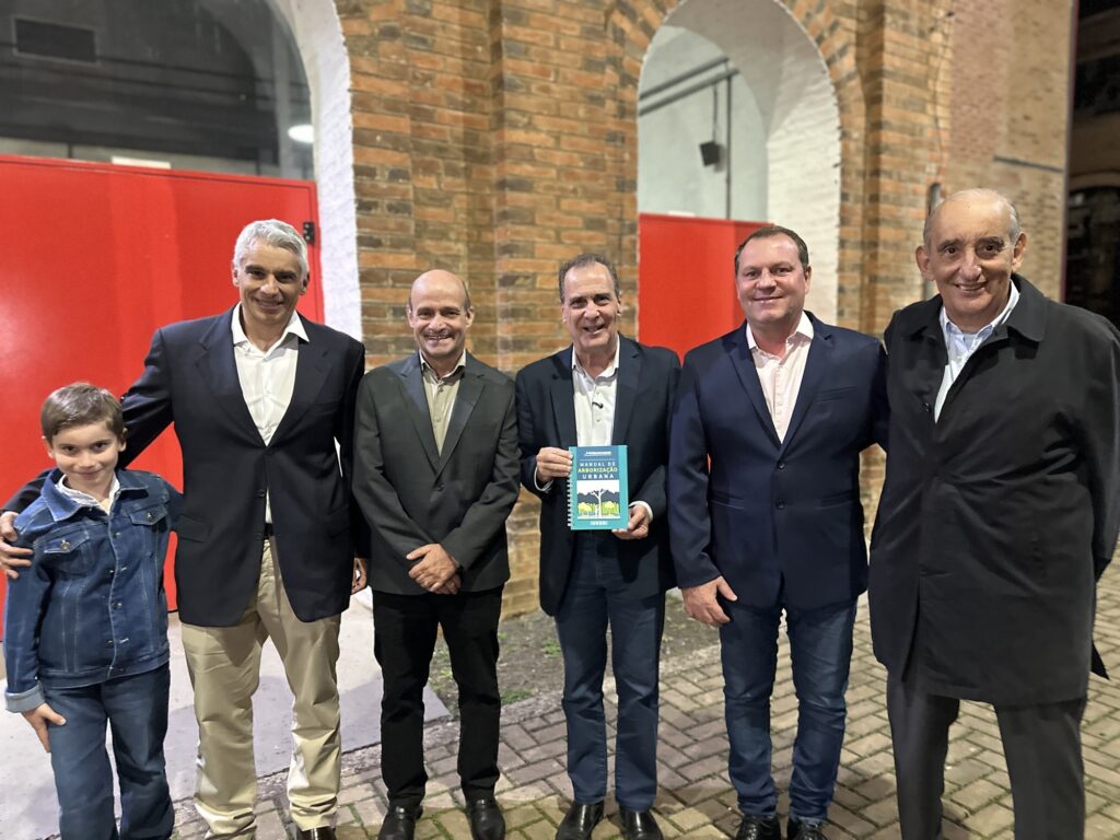 Foto de um grupo de pessoas posando em frente a uma fachada de tijolos com arcos — provavelmente em evento institucional ou lançamento. Observações detalhadas: Composição: seis adultos em pé alinhados no centro da imagem, com um garoto à esquerda parcialmente incluído; todos virados para a câmera e sorrindo. Idades e gênero: adultos (aparência masculina) e uma criança (menino) em primeiro plano à esquerda. Não faço identificação de pessoas. Roupas e aparência: a maioria veste paletós ou blazers escuros sobre camisas claras; um homem usa sobretudo preto; o garoto veste jaqueta jeans e camiseta. Trajes indicam ocasião formal/semiforma. Ação/objeto: o homem no centro segura um livro/caderno azul com o título legível “MANUAL DE ARBORIZAÇÃO URBANA” — parece ser o objeto do evento (lançamento ou apresentação). Expressões: sorrisos e postura relaxada, atmosfera cordial e comemorativa. Cenário/arquitetura: parede de tijolos expostos com arcos e portas/vermelhas ao fundo; piso de pedras/pavers sob os pés — ambiente remete a prédio histórico ou espaço cultural. Iluminação: luz artificial forte sobre os rostos, cores vivas (vermelho das portas ao fundo contrasta com o tijolo). Fotos tirada à noite ou em ambiente interno com iluminação dirigida. Detalhes adicionais: todos calçam sapatos sociais; há pequenas áreas de sombra nas laterais; enquadramento horizontal capturou do peito para baixo parcialmente (pernas e sapatos visíveis), e a imagem tem leve desfoque de profundidade nas extremidades.