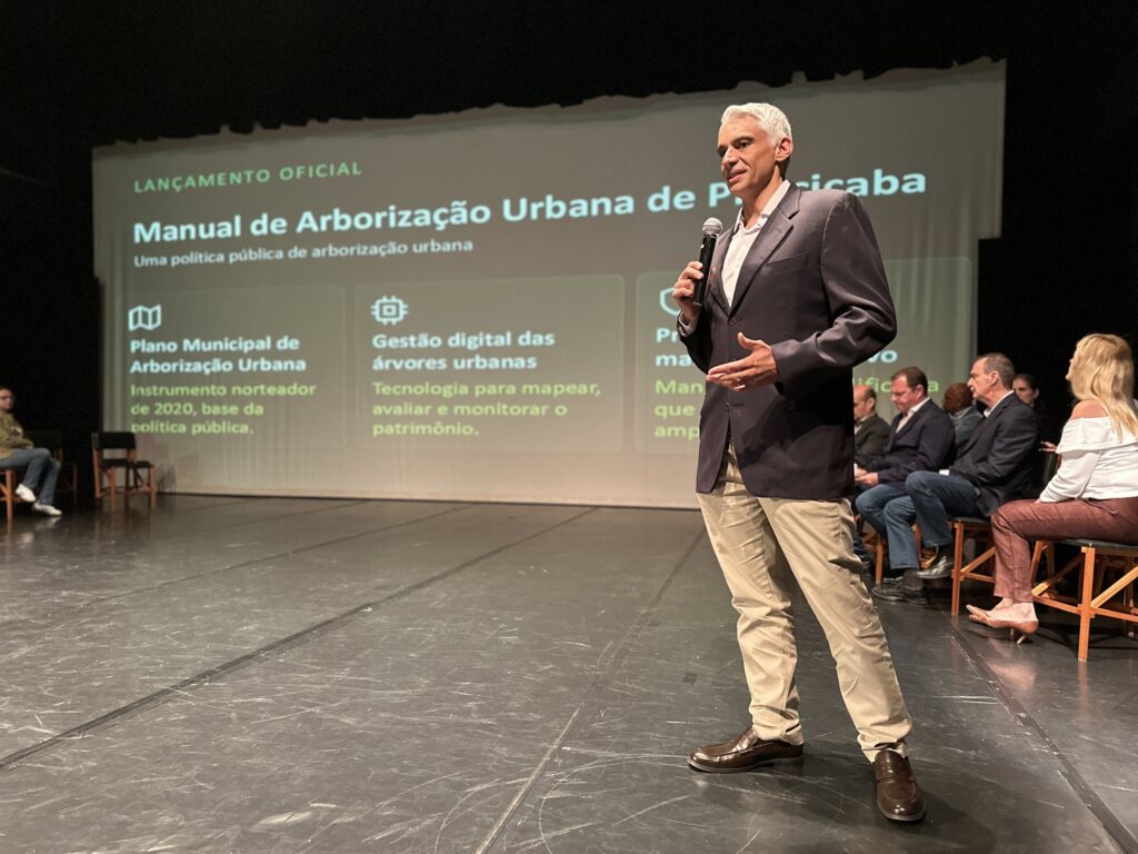 Foto de um evento institucional no palco: um orador em primeiro plano fala ao microfone diante de uma projeção que anuncia o "Manual de Arborização Urbana de Piracicaba". Observações principais: Orador: homem adulto em traje social (paletó escuro, camisa clara, calça bege), segurando microfone e gesticulando enquanto fala. Não faço identificação de pessoas. Projeção: tela grande ao fundo com o título "Manual de Arborização Urbana de Piracicaba" e blocos de texto/ícones sobre temas como plano municipal e gestão digital de árvores. No topo lê‑se "LANÇAMENTO OFICIAL". Cenário: palco de auditório com piso escuro; várias pessoas sentadas em cadeiras ao fundo, formando uma mesa/painel. À direita, parte do público/participantes visíveis de perfil. Iluminação: luz de palco direcionada no orador; fundo levemente desfocado, realçando quem fala. Contraste entre área iluminada e cortina/parede escura ao redor. Composição/fotografia: enquadramento horizontal com foco no orador à direita; profundidade que mostra tanto o público no palco quanto a projeção informativa. Atmosfera: formal e institucional, ligada a políticas públicas de arborização/planejamento urbano.