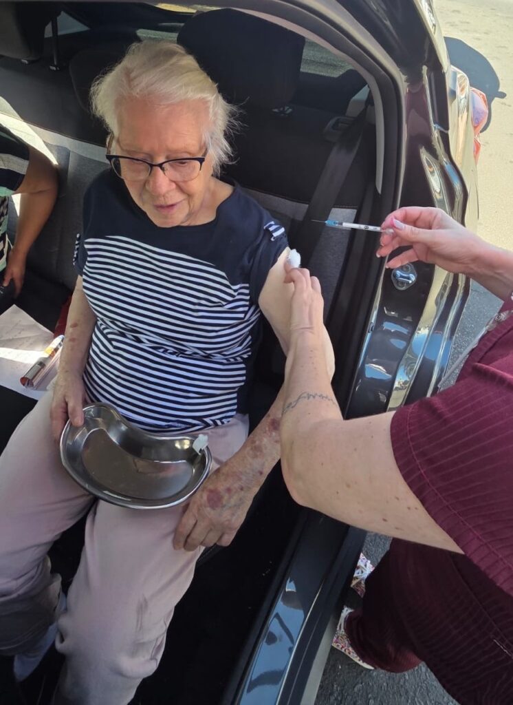 Nesta imagem, uma senhora idosa está sentada no banco traseiro de um carro enquanto recebe uma vacina ou injeção. Aqui estão os detalhes: A senhora: Ela tem cabelos brancos, usa óculos de armação escura, uma blusa com listras horizontais azul-marinho e brancas, e calças claras. Ela segura uma bandeja metálica (tipo rim) no colo. O procedimento: Uma profissional de saúde, que veste uma blusa de cor bordô, está aplicando a injeção no braço esquerdo da senhora. A profissional segura a seringa com uma mão e usa um algodão com a outra para preparar ou pressionar o local da aplicação. Ambiente: A cena acontece dentro do carro, com a porta aberta, indicando uma ação de vacinação no sistema drive-thru ou atendimento domiciliar/móvel.