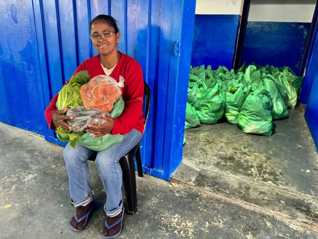 mulher sentada em uma cadeira plástica preta junto a uma porta/parede azul; segura várias sacolas e alimentos frescos no colo. À direita, no interior do cômodo aberto, há muitas sacolas verdes empilhadas no chão — parece um local de armazenamento ou distribuição de cestas alimentares. Pessoa: mulher adulta, pele morena, usa óculos, sorrindo de forma calma e acolhedora; cabelo preso para trás. Veste suéter vermelho sobre camiseta clara, calça jeans e chinelos com meia. Não é possível identificar quem é. Alimentos e embalagens: no colo dela há folhas verdes grandes (provavelmente couve ou repolho), um saco plástico com cenouras visíveis e outros itens embalados; as sacolas verdes ao fundo aparentam conter hortaliças semelhantes. Ambiente: piso de concreto gasto com marcas de uso; paredes e portas pintadas de azul intenso. O espaço tem aparência de depósito comunitário, cozinha social ou ponto de entrega de alimentos. Tonalidade e sensação: cores vivas (azul do fundo, vermelho do suéter, verde das verduras) e composição transmitindo cuidado, organização e solidariedade; atmosfera acolhedora e de trabalho comunitário.