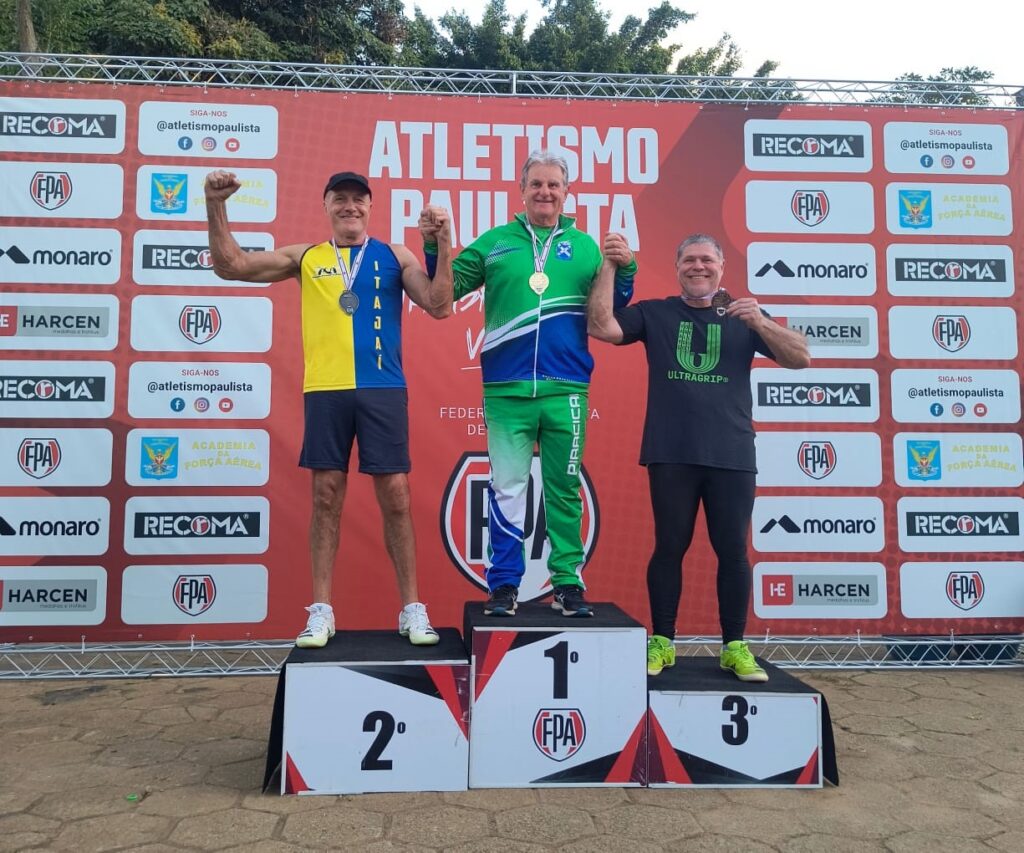 Uma foto que mostra o pódio de uma competição de atletismo organizada pela Federação Paulista de Atletismo (FPA). Três atletas estão posicionados sobre os blocos de premiação: 1º Lugar (Centro): Um homem vestindo um conjunto esportivo verde, azul e branco. Ele usa uma medalha de ouro no pescoço e tem a palavra "PIRACICABA" estampada na lateral da calça. Ele está sorrindo e erguendo o braço do atleta à direita. 2º Lugar (Esquerda): Um atleta vestindo uma camisa regata amarela e azul com a palavra "ITAJAÍ" escrita na vertical, e shorts azul marinho. Ele usa uma medalha de prata e está com o braço erguido em sinal de comemoração. 3º Lugar (Direita): Um atleta vestindo uma camiseta preta com a estampa verde "ULTRAGRIP" e calças pretas. Ele segura sua medalha de bronze e sorri para a foto. Ao fundo, há um painel promocional da FPA (Federação Paulista de Atletismo) com várias marcas parceiras (como RECOMA, Monaro, Harcen) e os logos da Academia da Força Aérea. O chão é pavimentado com pedras irregulares.
