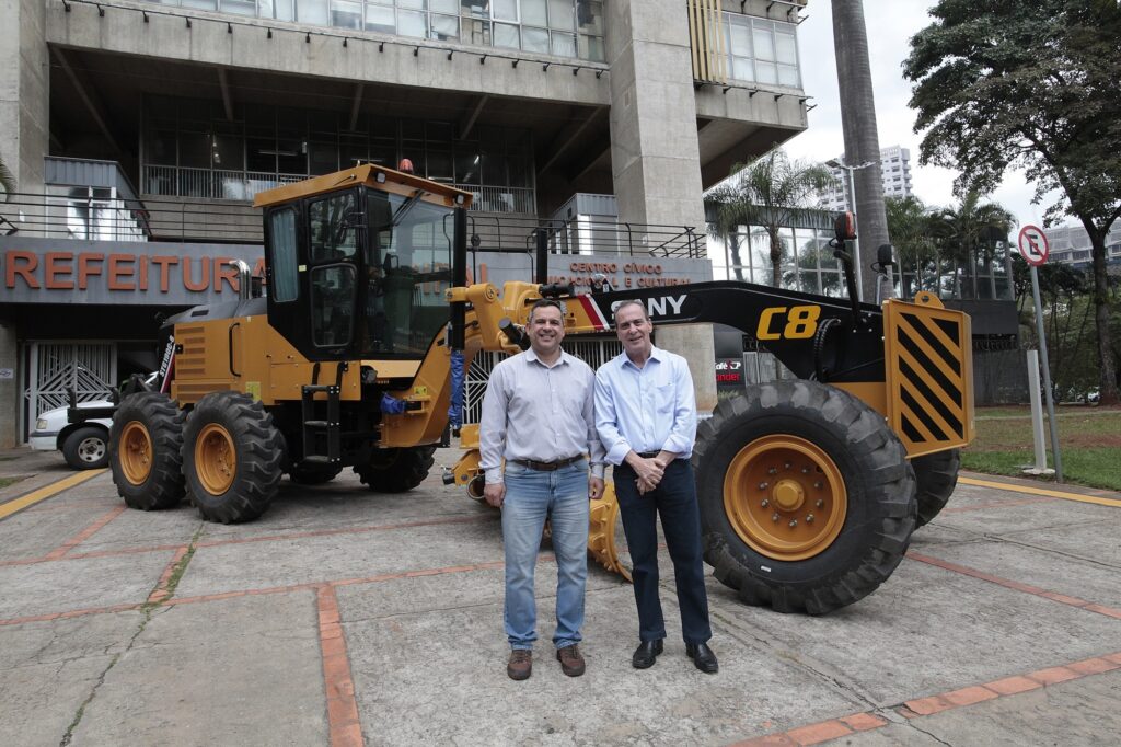 Uma foto externa mostrando duas pessoas em primeiro plano na frente de um veículo pesado de obras (motoconformadora) amarelo. Pessoas: dois homens de pé lado a lado, sorrindo. Ambos usam camisa social (um de manga comprida azul-claro e outro de manga comprida branca com riscas finas) e calças. Não vou tentar identificar quem são. Veículo: uma motoconformadora/amarelo com grandes pneus traseiros, braços hidráulicos e cabine envidraçada; na lateral aparece a letra "C8". Cenário: em frente a um prédio de concreto com a fachada e letras parcialmente visíveis indicando "PREFEITURA" e "CENTRO CÍVICO"; há árvores e postes ao fundo. Piso: calçada/pátio de concreto com marcações em tijolo/cerâmica formando quadrados. Clima e luz: dia nublado, iluminação natural difusa. Atmosfera: parece ser um evento oficial ou entrega de equipamento relacionado a obras públicas.
