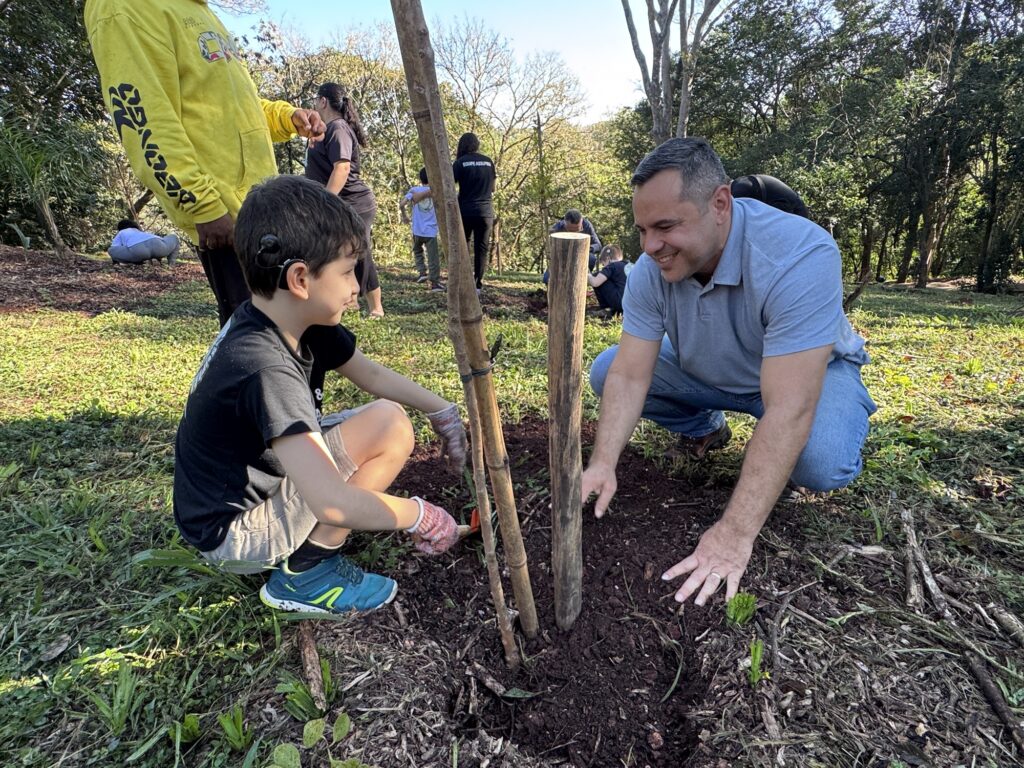 Esta imagem registra um momento de conexão e educação ambiental, mostrando um adulto e uma criança plantando uma árvore juntos em uma área arborizada. A Ação Principal: Um homem adulto (vestindo camisa polo cinza e jeans) e um menino (de camiseta preta e bermuda) estão agachados na terra, focados no plantio. O menino, que usa um aparelho auditivo, está manuseando uma pequena ferramenta de jardinagem, enquanto o homem ajuda a ajeitar a terra ao redor da base da muda, que está apoiada por uma estaca de bambu. O Ambiente: É um local ensolarado e ao ar livre, com um gramado coberto por folhas secas e terra. O cenário sugere um parque ou uma área de preservação. Contexto: Ao fundo, vemos outras pessoas participando da mesma atividade, algumas também agachadas cuidando de outras mudas. Uma pessoa de moletom amarelo está em pé próxima ao grupo principal, e outras vestem camisetas pretas (algumas com a inscrição "EQUIPE ASSUPRA"), indicando que se trata de uma ação coletiva ou mutirão de plantio. Clima: A cena transmite cuidado, colaboração e o aprendizado prático sobre preservação da natureza. O sorriso do homem e a concentração da criança destacam o aspecto positivo desse momento.