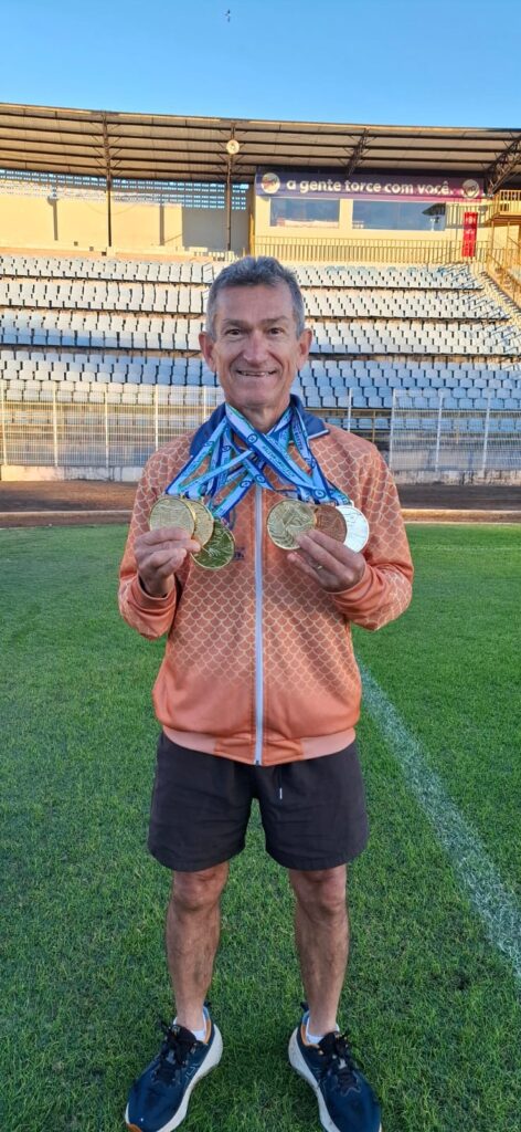 Foto de um homem adulto mais velho sorrindo num campo/estádio, segurando várias medalhas. Principais elementos: Pessoa: um homem de aproximadamente 50–70 anos, sorrindo para a câmera; não posso identificar ou nomear. Medals: várias medalhas (ouro, prata e bronze aparentes) penduradas no pescoço e seguradas com as mãos à frente, com fitas em tons de azul e branco. Roupa: jaqueta esportiva laranja com padrão em forma de escamas, bermuda escura e tênis esportivos. Local: campo gramado com linhas de marcação; ao fundo, arquibancada vazia e estrutura coberta. No topo da arquibancada há um banner com o texto “a gente torce com você”. Atmosfera: clima de comemoração e conquista — a expressão do rosto e as medalhas indicam orgulho por resultados esportivos ou participação em competição. Detalhes menores: céu limpo e luz suave de fim de tarde; piso bem cuidado; cercado e gradis entre campo e arquibancada.