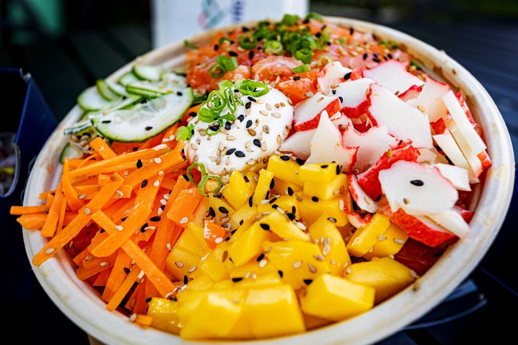 Bowl colorido visto de perto, preenchido com ingredientes frescos e cortados em cubos/tiras. Detalhes observáveis: Componentes principais: cubos de manga amarela brilhante; tiras finas de cenoura laranja; fatias finas de pepino ao fundo; fatias brancas com borda avermelhada que parecem kani (imitação de caranguejo); pedaços alaranjados no fundo que sugerem salmão cru picado. Centro cremoso: uma porção de molho ou creme branco (parece maionese temperada ou molho à base de iogurte/aji) coberta por sementes de gergelim e cebolinha fatiada. Temperos e finalização: sementes de gergelim preto e branco espalhadas por toda a superfície; cebolinha verde picada como toque final. Texturas e cores: contraste entre a maciez da manga e do peixe, a crocância da cenoura e do pepino, e a cremosidade do molho; paleta vibrante — amarelos, laranjas, verdes e branco-avermelhado. Apresentação: servido em bowl descartável/biodegradável, visual muito atraente e típico de poke/salad bowl japonês-havaiano moderno. Atmosfera sugerida: refeição fresca, leve e refrescante, adequada para almoço rápido ou refeição saudável.