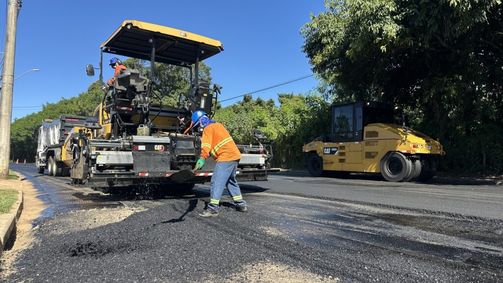 Esta imagem mostra uma cena de obra de pavimentação asfáltica em uma rua durante um dia ensolarado. Equipamentos: Há uma grande vibroacabadora da marca CAT (Caterpillar) em ação, espalhando o asfalto quente. Atrás dela, um pouco mais adiante na via, está um rolo compactador amarelo, também da marca CAT, pronto para finalizar a compactação do material. Trabalhadores: Um operário, vestindo uniforme laranja de alta visibilidade, calça jeans, capacete azul e máscara de proteção, está em primeiro plano usando uma pá para organizar manualmente a massa asfáltica. Outro operário está operando a máquina principal. Ambiente: O céu está limpo e azul. Há vegetação ao fundo e o asfalto recém-aplicado, ainda escuro e brilhante, cobre parte da via. Também é visível um caminhão basculante branco aguardando próximo à vibroacabadora.