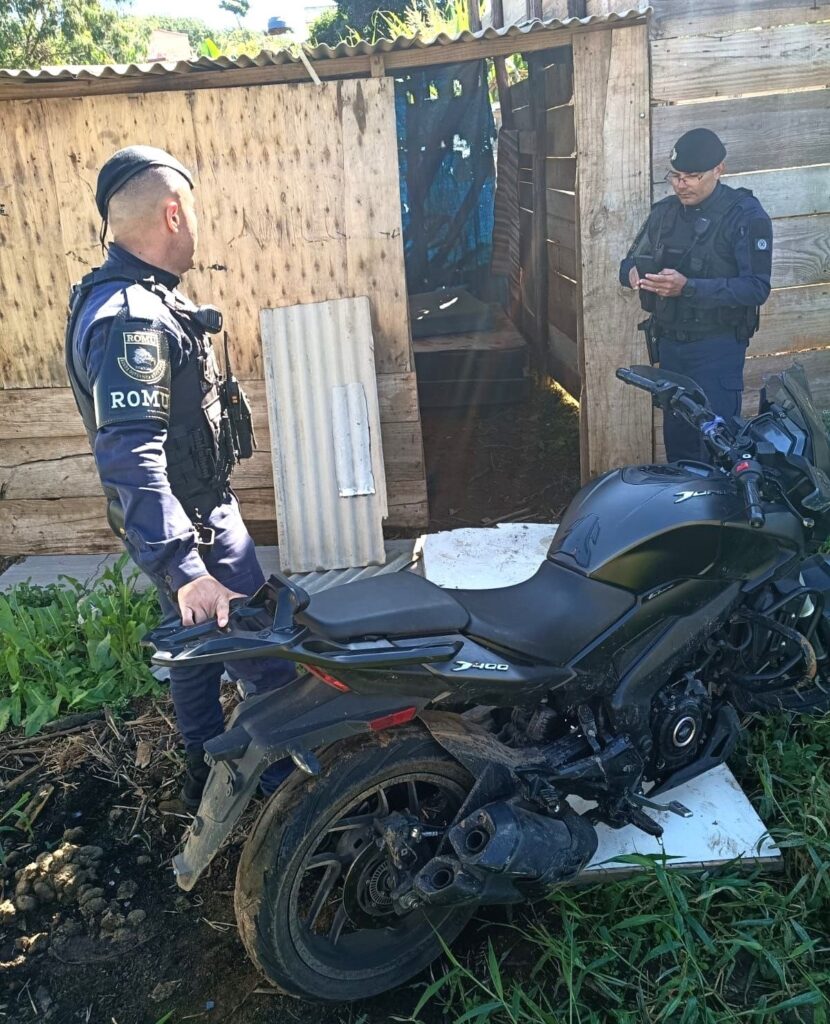 Foto de uma abordagem/ocorrência externa com policiais e uma motocicleta preta em primeiro plano. Cena geral: dois agentes fardados (coletes táticos, distintivo/palavra “ROMU” visível na manga de um deles) em frente a uma construção rústica de madeira com uma porta aberta que dá para um interior pouco iluminado. Ação: um policial à esquerda segura a parte traseira da motocicleta; o outro, à direita, consulta um aparelho (provavelmente celular) enquanto está junto à entrada da construção. Motocicleta: modelo esportivo/prestador urbano de cor preta (no tanque lê-se "J400"), com sujeira e lama nas rodas e no escapamento, indicando que esteve em terreno enlameado ou sofreu queda; aparenta danos leves/respingos na parte inferior do motor e no para-lama traseiro. Ambiente: solo de terra e vegetação rasteira ao redor; placa de fibrocimento encostada na porta; iluminação de dia com sombras fortes sugerindo sol alto. Detalhes visuais: acesso improvisado à construção (madeiras cruas, telha ondulada), pequenos detritos no chão e rastro de terra ao redor da motocicleta.