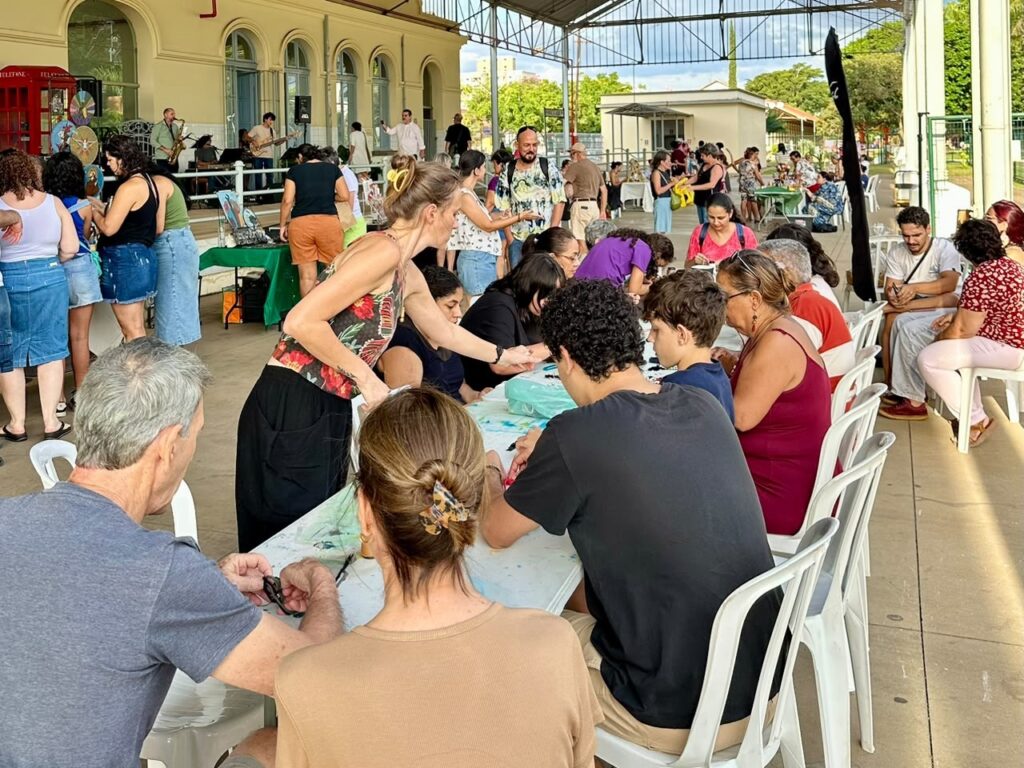 Esta imagem registra uma atividade coletiva ao ar livre, possivelmente uma oficina de artesanato ou artes manuais, acontecendo em uma área coberta, como um pavilhão ou estação antiga. Atividade principal: Um grupo de pessoas está sentado em volta de longas mesas, concentrado em criar algo com as mãos. Uma instrutora, em pé, estende a mão para auxiliar um dos participantes. Há materiais espalhados sobre a mesa, sugerindo trabalhos manuais. Ambiente: O espaço é amplo e aberto, com um telhado metálico. Ao fundo, nota-se uma estrutura arquitetônica clássica, com janelas em arco. Detalhes adicionais: À esquerda, no fundo, há um pequeno grupo de músicos se apresentando (um saxofonista e um violonista), o que traz um ar de descontração ao evento. Há uma cabine telefônica vermelha em estilo retrô, compondo a decoração do cenário. O clima parece ser de uma tarde cultural ou recreativa, com várias pessoas circulando pelo local, observando outras barracas e aproveitando a movimentação.