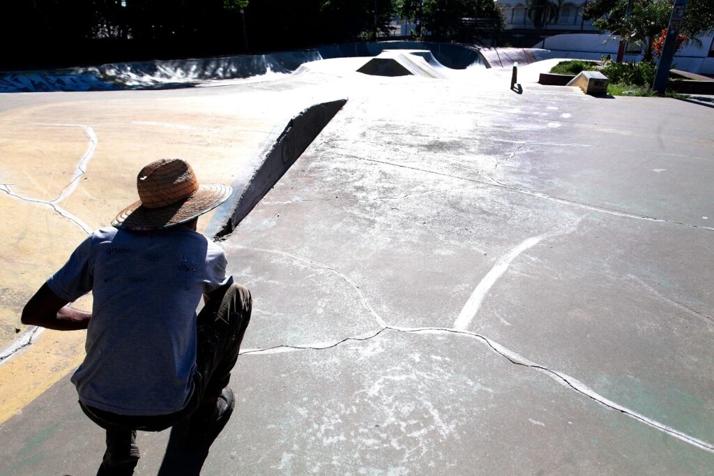 Um homem agachado de costas, vestindo camiseta cinza e chapéu de palha largo, ocupa o canto inferior esquerdo da foto. Ele parece observar ou preparar algo no chão de concreto de um skate park vazio — há rampas, transições e um pequeno corrimão ao fundo. O piso está gasto, com várias fissuras preenchidas por selante branco e manchas claras que brilham sob a luz forte do sol. Ao fundo, à direita, há um canteiro com plantas e algumas árvores que projetam sombra na borda do parque; ao longe vê-se uma estrutura branca (talvez muro ou prédio). A cena transmite calor, calmaria e a sensação de um lugar usado mas tranquilo naquele momento.