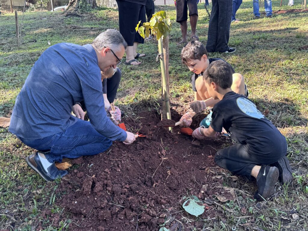 Esta imagem mostra um momento educativo e muito bonito de plantio de uma árvore. A Ação: Um adulto e duas crianças estão agachados na terra, trabalhando juntos para fixar uma muda de árvore. O adulto, de camisa azul e óculos, está do lado esquerdo, enquanto as duas crianças (um menino mais novo e outro um pouco maior) estão do lado direito. Eles estão usando pequenas pás manuais para mover a terra ao redor da base da muda, que está amparada por uma estaca de madeira. O Ambiente: É uma área externa ensolarada com gramado. Vemos os pés e as pernas de outras pessoas ao redor, sugerindo que é uma atividade em grupo, possivelmente um evento comunitário ou escolar de preservação ambiental. Os Detalhes: Todos parecem concentrados na tarefa. As crianças usam luvas de jardinagem e roupas confortáveis. A terra parece bem preparada e fértil, com uma cor avermelhada característica de muitas regiões do Brasil. É um registro inspirador de educação ambiental e colaboração intergeracional!