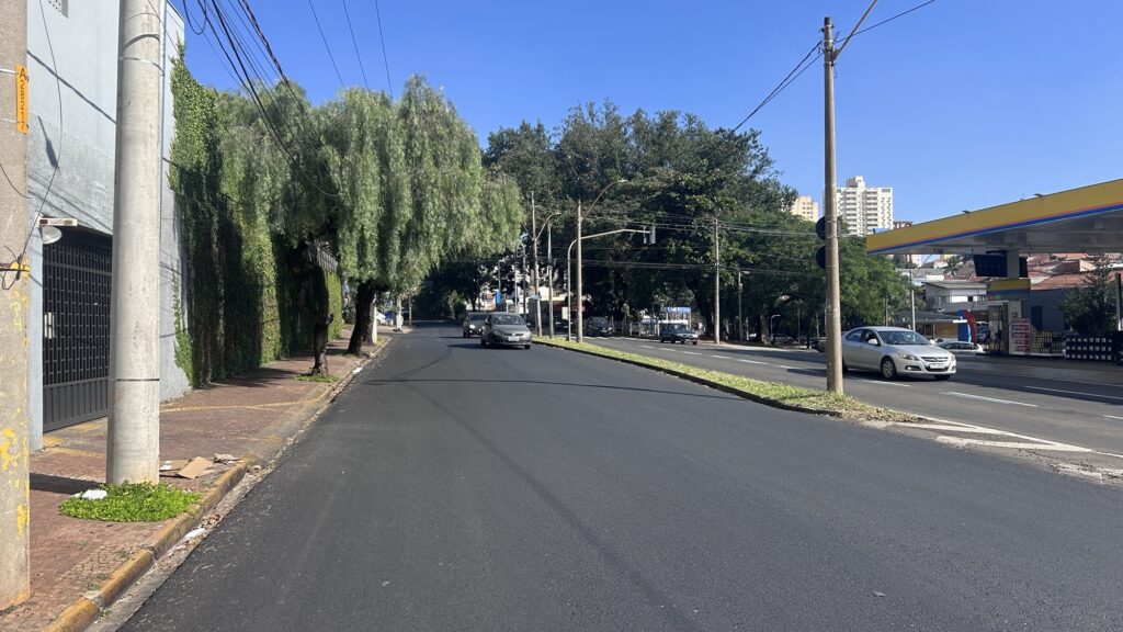 Rua larga em dia claro, vista em perspectiva para cima da ladeira. Pavimento: asfalto escuro e aparentemente novo cobre a pista principal; faixa central com canaleta gramada separa os sentidos. Tráfego: poucos carros circulando — um carro prata mais próximo vindo em direção ao observador e outros mais distantes. Calçada esquerda: passeio em piso de lajota, poste de concreto próximo à borda, caixas de papelão e lixo junto ao meio-fio; muro coberto por trepadeira verde. Vegetação: árvores de copa densa à esquerda e ao fundo, algumas com galhos pendentes que sombreiam a calçada. Postes e fiação: vários postes e muitos cabos elétricos cruzando a cena, visíveis contra o céu. Comércio/infraestrutura: posto de combustível à direita com cobertura amarela; prédios residenciais ao fundo mais à direita. Céu/clima: azul intenso, céu limpo — luz forte e sombras bem definidas, indicando manhã/tarde ensolarada. Atmosfera geral: cena urbana tranquila, parecendo uma rua residencial/comercial com obras recentes no pavimento.