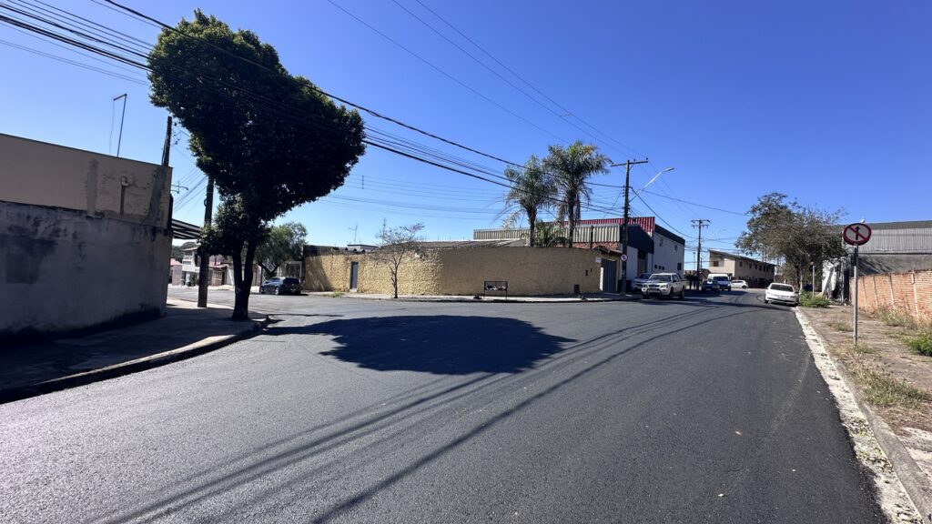 Na imagem, vemos uma rua de bairro sob céu azul e ensolarado, com o pavimento recém-finalizado e de aspecto liso e uniforme. À esquerda, destaca-se uma árvore de copa densa e irregular, próxima a uma construção de fachada desgastada. Ao fundo, aparecem casas e prédios de diferentes estilos, alguns com paredes coloridas e telhados vermelhos, compondo um cenário típico urbano. Do lado direito da via, há carros estacionados e um sinal de trânsito indicando a proibição de conversão à esquerda. Postes e fiação aérea acompanham toda a extensão da rua, reforçando o ambiente cotidiano de uma vizinhança residencial.