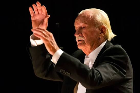 A imagem mostra um senhor mais velho, provavelmente um maestro, regendo.Detalhes:Pessoa: Homem idoso, com cabelos brancos e lisos penteados para trás e bigode branco. A expressão é concentrada e intensa, com a boca levemente aberta como se estivesse dando a entrada.Roupa: Terno preto, camisa branca e gravata clara. Visual clássico de maestro ou regente de orquestra/coral.Gesto: Está de lado, com os dois braços levantados. A mão direita está aberta, com a palma para frente, e a esquerda parece estar marcando o tempo. Transmite autoridade e emoção.Fundo: Preto, sem distrações. A luz está focada nele, destacando o rosto, as mãos e o terno. Isso dá um clima de palco e importância ao momento.A foto passa elegância, experiência e paixão pela música. Parece um registro de uma apresentação ao vivo.
