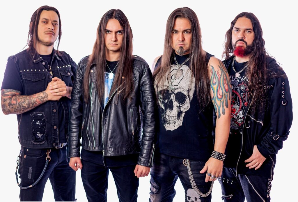 Esta é uma foto promocional de uma banda de heavy metal com quatro integrantes, posando lado a lado contra um fundo branco. Da esquerda para a direita: O primeiro membro tem cabelos longos e escuros, usa um colete preto sem mangas com tachas nos ombros e braços tatuados. O segundo membro veste uma jaqueta de couro preta, tem cabelos lisos longos e mantém uma expressão séria. O terceiro membro, no centro, também de cabelos longos, veste uma camiseta preta com uma estampa grande de caveira no peito e possui uma tatuagem colorida de chamas no braço esquerdo. O quarto membro, à direita, tem uma barba com detalhes em vermelho, cabelos longos e veste uma camiseta preta com estampas geométricas e detalhes com ilhoses. O visual de todos é bem característico do estilo metal, com roupas pretas, correntes e uma postura imponente.