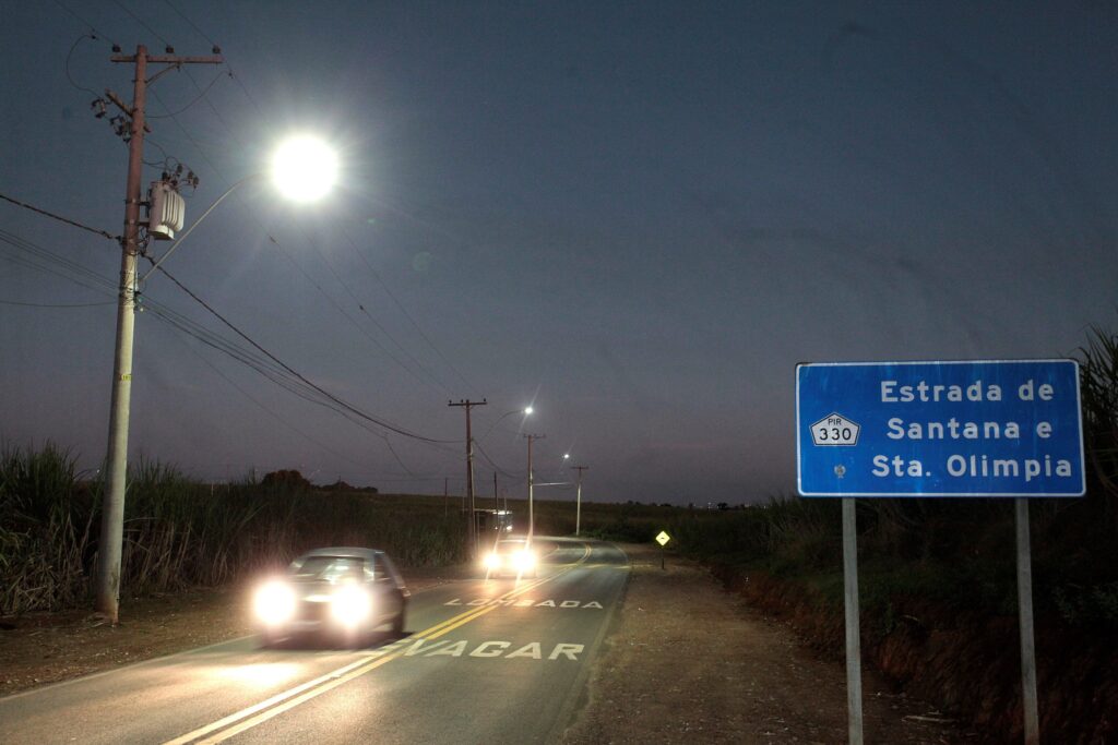Trecho de estrada rural à noite/crepúsculo, fotografado em perspectiva que aponta para baixo pela via. Cena com iluminação pública intensa e alguns carros em movimento com faróis acesos. Sinalização e marcações Placa azul à direita com texto parcialmente legível: “Estrada de Santana e Sta. Olímpia” e o símbolo PIR 330. Pintura no asfalto no primeiro plano com as palavras “DEVAGAR” e “LOMBADA”, indicando aviso de redução de velocidade. Iluminação e infraestrutura Poste de energia/transformador em primeiro plano à esquerda; luminária de rua muito brilhante produz flare na imagem. Série de postes ao longo da estrada criando pontos de luz que guiam a linha da via até o horizonte. Fiação elétrica visível conectando os postes. Tráfego e movimento Pelo menos dois veículos em movimento na pista, faróis brilhantes causando reflexos e leve efeito de borrão por movimento. Estrada de pista simples com faixa dupla amarela central. Ambiente e cenário Laterais da via dominadas por vegetação alta — aparenta ser canavial ou plantação — e taludes de terra
