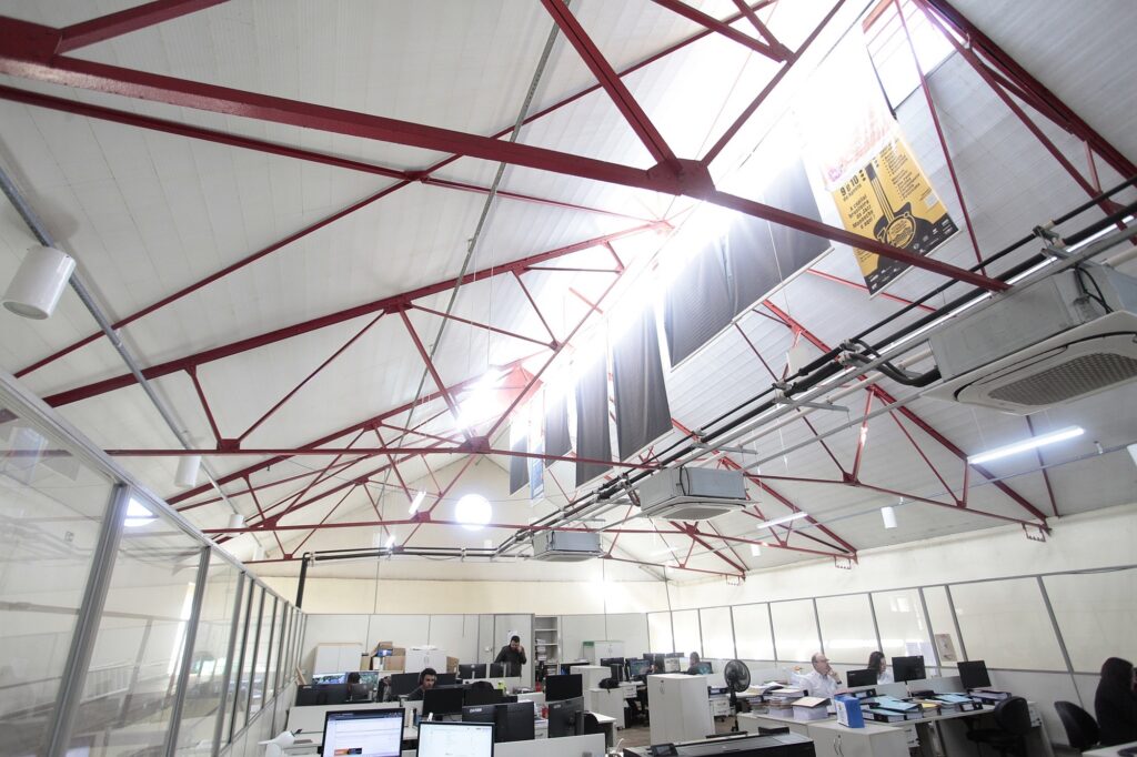 Esta é uma foto de um ambiente de trabalho moderno e amplo, com um estilo industrial bem marcante. Aqui estão os pontos principais: O Ambiente: O teto é alto, com vigas de metal vermelhas aparentes que cruzam todo o espaço, dando um visual estruturado. Há uma entrada de luz natural vindo de cima, o que ilumina bem o local. Decoração: Pendurado no teto, há um pôster informativo grande, com fundo amarelo, que parece divulgar um evento ou festival (com referências a música e datas). Estrutura: O escritório é organizado em formato de "ilha", com divisórias de vidro e estações de trabalho brancas. As mesas estão equipadas com computadores, monitores e documentos. As Pessoas: Várias pessoas estão sentadas em suas mesas, concentradas trabalhando em seus computadores. Algumas estão em grupos de trabalho e outras sozinhas. Clima: É um escritório que parece bem funcional e colaborativo, com uma boa dose de luz natural e um visual que mistura a praticidade de um local de trabalho com uma arquitetura bem característica.