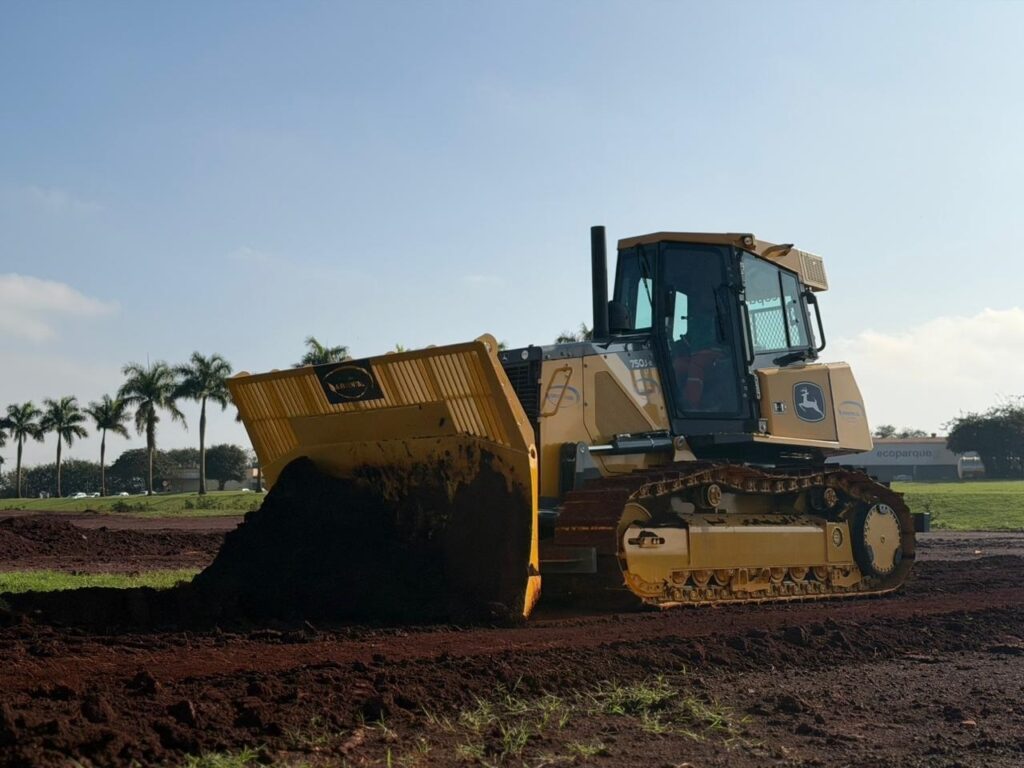 Foto de um trator de esteiras (bulldozer) amarelo trabalhando em um terreno de terra avermelhada. Máquina: trator de esteiras com cabine fechada; marca aparente (símbolo) na lateral. Ação: lâmina frontal inclinada empurra e alisa montes de terra vermelha; há terra aderida à lâmina e às esteiras. Perspectiva: tomada lateral-baixa, destacando a lâmina à esquerda em primeiro plano e as esteiras à direita. Solo e marcas: solo argiloso de cor vermelho-escuro com sulcos frescos deixados pelas esteiras. Ambiente: céu azul claro, poucas nuvens; vegetação baixa ao fundo e área aberta, possivelmente campo ou canteiro de obras. Iluminação: luz do dia forte, sombras suaves indicando manhã ou fim de tarde.