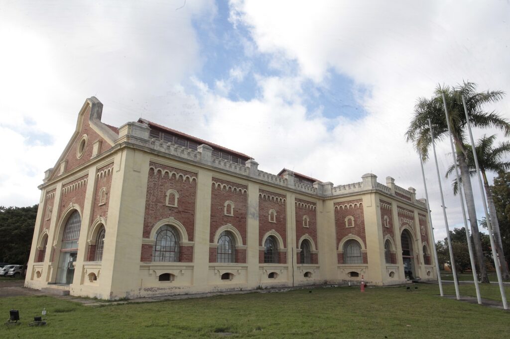 Esta é uma foto de um prédio histórico imponente, construído em tijolos aparentes avermelhados com molduras em amarelo claro. Aqui estão os detalhes: Arquitetura: O estilo é clássico e robusto, lembrando antigas instalações industriais ou de serviços públicos do final do século XIX ou início do século XX. Possui grandes janelas em arco com molduras decoradas e um telhado com telhas de barro. Identificação: Na lateral direita da fachada, há uma inscrição na parede onde se lê claramente a palavra "MATADOURO". O Entorno: O prédio está em um terreno gramado, com um céu azul parcialmente nublado ao fundo. Ao lado da construção, há quatro postes altos e finos que parecem ser mastros para bandeiras, alinhados verticalmente, e algumas palmeiras mais ao fundo. Estado: O edifício parece estar bem conservado, mantendo suas características originais de época, e possui portas de vidro modernas na entrada principal, indicando que pode ter sido restaurado ou adaptado para novos usos, como um centro cultural ou museu.