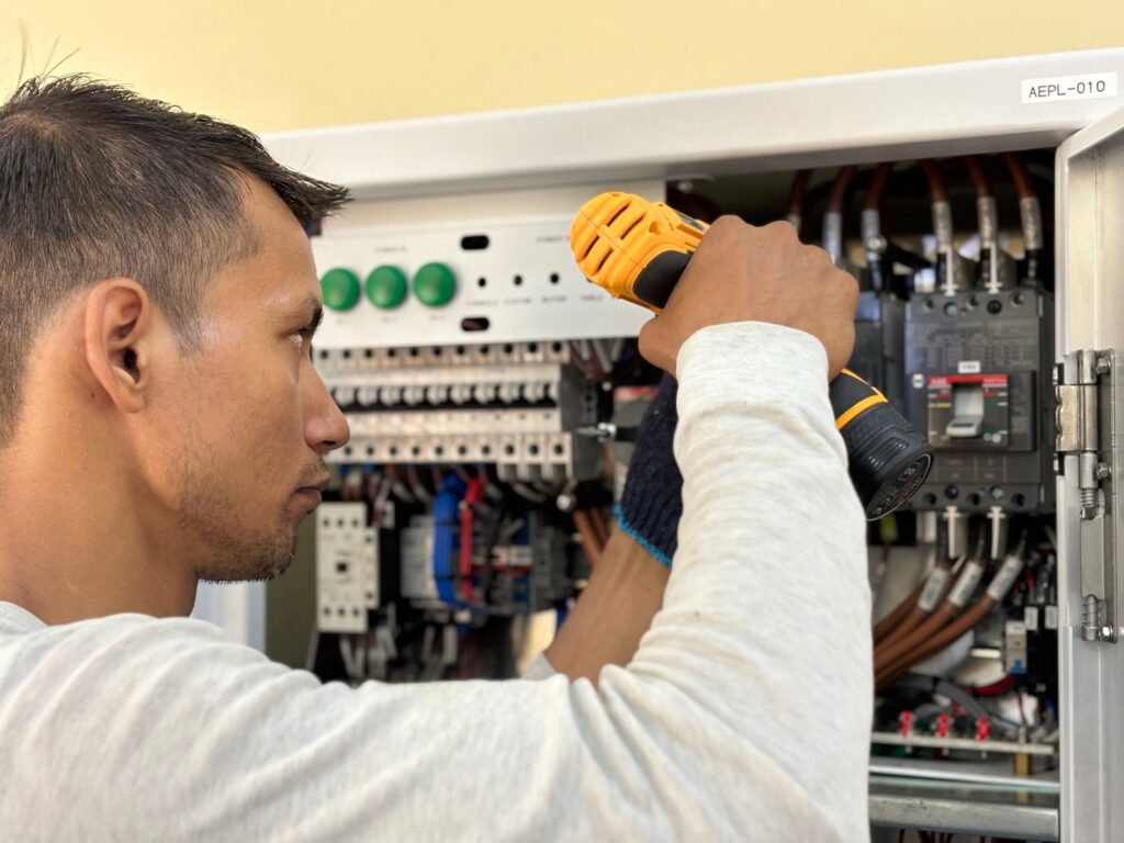 Foto colorida, horizontal, mostrando um homem trabalhando num quadro elétrico/centro de distribuição. A imagem é enquadrada em close lateral: o rosto do trabalhador aparece de perfil à esquerda (só parte da cabeça e do rosto), e as mãos dele, à direita, seguram uma parafusadeira elétrica amarela enquanto realiza um ajuste dentro do painel. Detalhes visuais: Pessoa: homem de pele parda, cabelo curto cortado rente nas laterais; apenas o perfil e parte do pescoço aparecem. Não é possível identificar quem é. Ação: ele usa a mão direita para segurar uma parafusadeira/cordão de ferramenta elétrica amarela e preta, apontada para dentro do painel. A mão esquerda segura ou estabiliza algo no interior do quadro; a manga é clara (tom bege/acinzentado). Painel elétrico: porta metálica aberta à direita revela vários componentes — disjuntores, bornes, cabos grossos de cor marrom com etiquetagem, conjuntos de conexões e um conjunto de blocos terminal/contatos. Ao fundo há um painel branco com três botões redondos verdes e várias fileiras de terminais/peças. Cabos e componentes: fios organizados, alguns com identificadores; cabos azuis e marrons visíveis; componentes eletrônicos e etiquetas pequenas, sugerindo instalação profissional e manutenção em equipamento de média/alta tensão ou distribuição. Etiqueta: no fechamento superior da caixa há uma etiqueta com o texto “AEPL–010”. Iluminação e foco: luz natural/ambiente clara; foco nítido no rosto e na ferramenta, com o interior do painel ligeiramente desfocado, criando profundidade de campo. Tom e sensação: cena de trabalho técnico, atenta e profissional — transmite concentração e cuidado na realização de manutenção ou instalação elétrica.