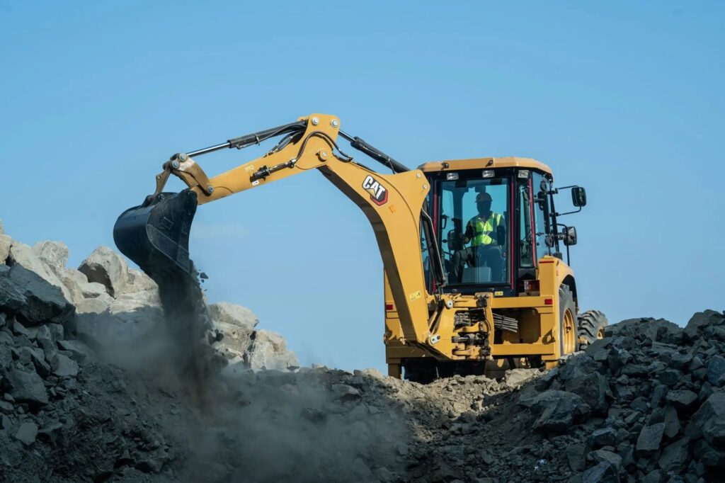 Nesta imagem, vemos uma retroescavadeira da marca CAT (Caterpillar) em operação. A Máquina: É um equipamento de construção robusto, de cor amarela característica, com uma caçamba frontal que está despejando terra ou pedras em um amontoado. O braço mecânico está estendido para o lado. O Operador: Dentro da cabine, que possui janelas de vidro, é possível ver o operador usando um colete refletivo de segurança, concentrado no controle da máquina. O Ambiente: A cena ocorre em um canteiro de obras ou área de mineração, onde há uma grande quantidade de rochas e terra movimentadas. O céu ao fundo está limpo e azul, indicando um dia ensolarado.