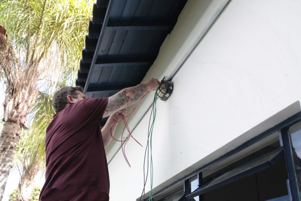 A imagem mostra um homem realizando um serviço de manutenção elétrica na parte externa de uma construção. Aqui estão os detalhes principais: O homem: Ele veste uma camiseta vinho e tem várias tatuagens no braço. Está concentrado, manipulando alguns fios de cores diferentes (vermelhos, verdes e pretos) que estão conectados a uma luminária de parede. A atividade: O foco do trabalho é uma luminária de estilo industrial, com uma grade de proteção, fixada na parede branca. Ele parece estar fazendo uma instalação ou reparo na fiação. O ambiente: A cena acontece ao ar livre, sob o beiral escuro de um telhado. Ao fundo, à esquerda, é possível ver a copa de uma palmeira alta, indicando que o local é uma área externa.