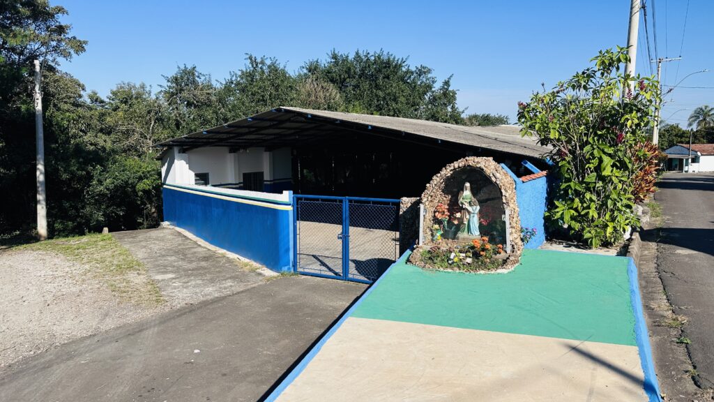 Vista externa de um pequeno estabelecimento pintado de azul com grade e portão na entrada; à direita, uma gruta artesanal emoldura uma imagem religiosa (Nossa Senhora) rodeada de flores e vasos. O prédio tem cobertura metálica inclinada e parede branca no interior; atrás dele há árvores densas com folhagem verde e céu limpo azul indicando dia ensolarado. À direita da cena há uma faixa de calçada pintada de verde, um canteiro com arbustos altos e postes de luz ao longo da rua tranquila — a composição transmite ambiente urbano periférico, calmo e bem cuidado.