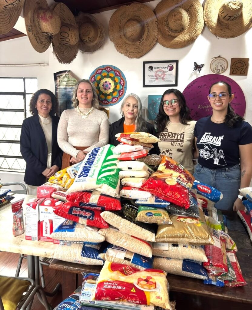Esta foto mostra cinco mulheres reunidas atrás de uma grande mesa cheia de alimentos, o que sugere uma ação de arrecadação de doações ou uma campanha solidária. Aqui estão os detalhes principais: As pessoas: São cinco mulheres de diferentes idades posando para a foto. Elas estão sorrindo e parecem estar em um ambiente interno, possivelmente um centro comunitário ou escritório. A doação: Sobre a mesa, há uma pilha considerável de alimentos não perecíveis, incluindo muitos pacotes de arroz de várias marcas, feijão, farinha de milho e caixas de leite. O ambiente: A parede ao fundo está decorada com itens artesanais e informativos: Há vários chapéus de palha pendurados no alto. Uma mandala colorida feita à mão. Um certificado em uma moldura preta. Um círculo de madeira com inscrições (parece ser decorativo ou comemorativo). Duas das mulheres estão vestindo camisetas temáticas de "Arraiá", indicando que pode ser uma iniciativa ligada a festas juninas ou eventos de arrecadação sazonal. A cena transmite um sentimento de solidariedade e trabalho em equipe voltado para o bem comum.